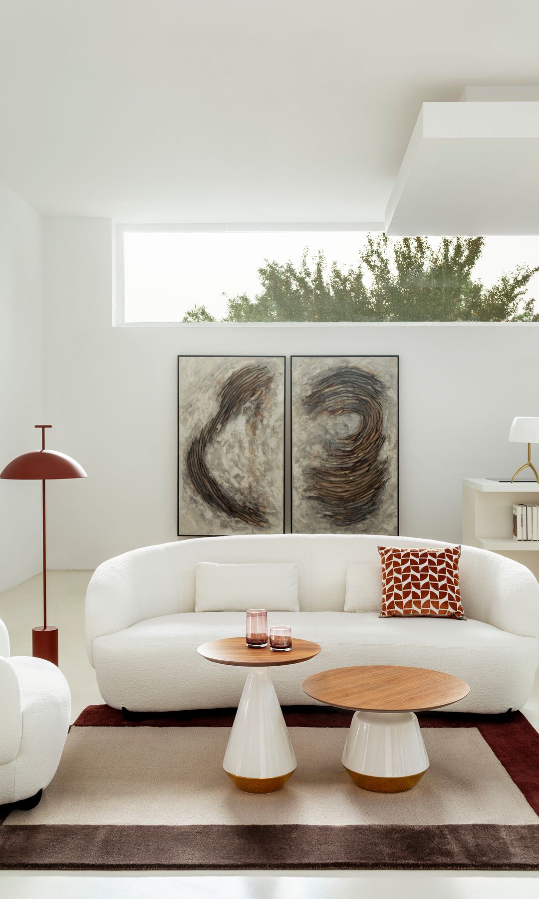 Salón con sofá blanco curvo, dos alfombras, cuadros en la pared, lámpara de pie roja, cojín rojo, mesas de centro con el sobre de madera y el pie rojo y blanco, sillón blanco