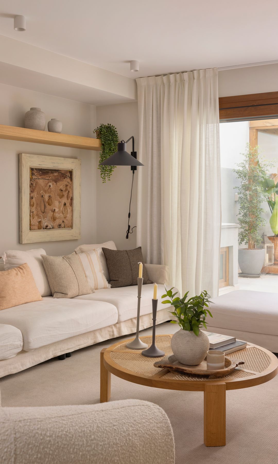 Salón en tonos beiges con salida a la terraza, sofá blanco, mesa de centro de madera, velas, jarrón, balda de madera, cuadro