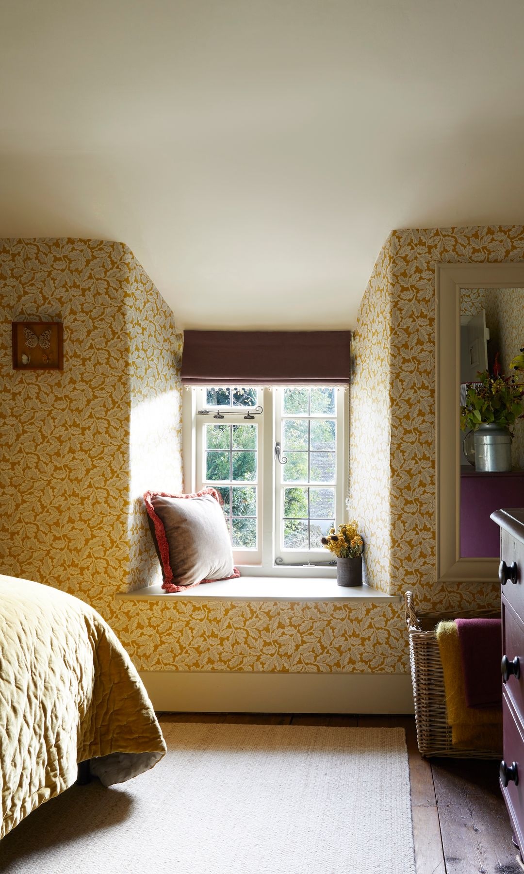 Dormitorio con papel pintado de flores amarillas, banco en el hueco de la ventana, cómoda oscura, cojines, estor, jarrón