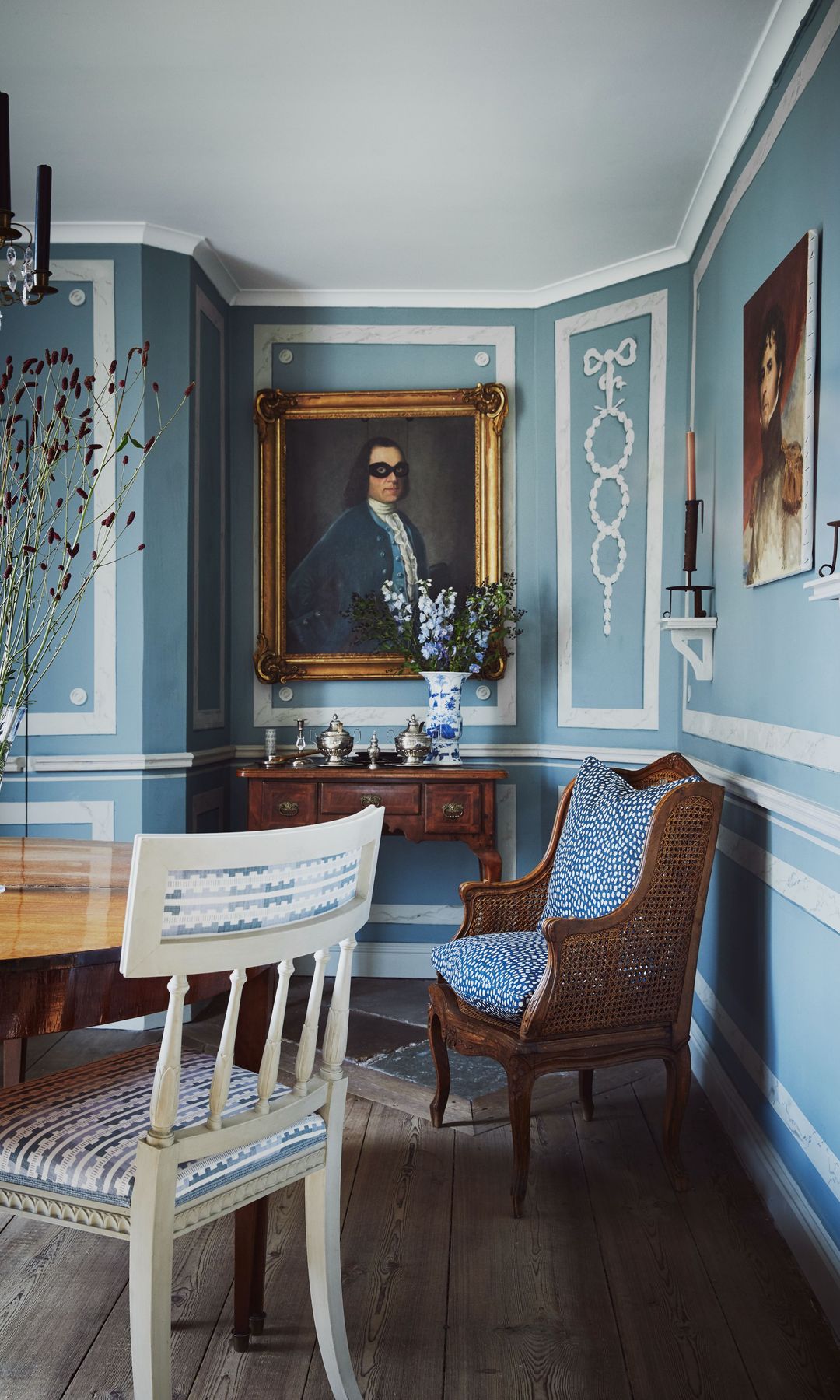Comedor con paredes azules con molduras, aparador antiguo, cuadros con el marco dorado y butaca de rejilla