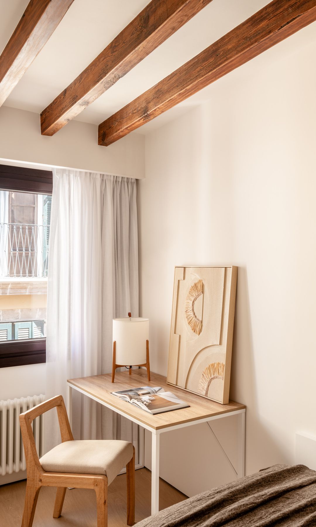 Rincón de escritorio junto a la ventana, vigas de madera, mesa de madera, cortinas, radiador, cuadro y lámpara de sobremesa