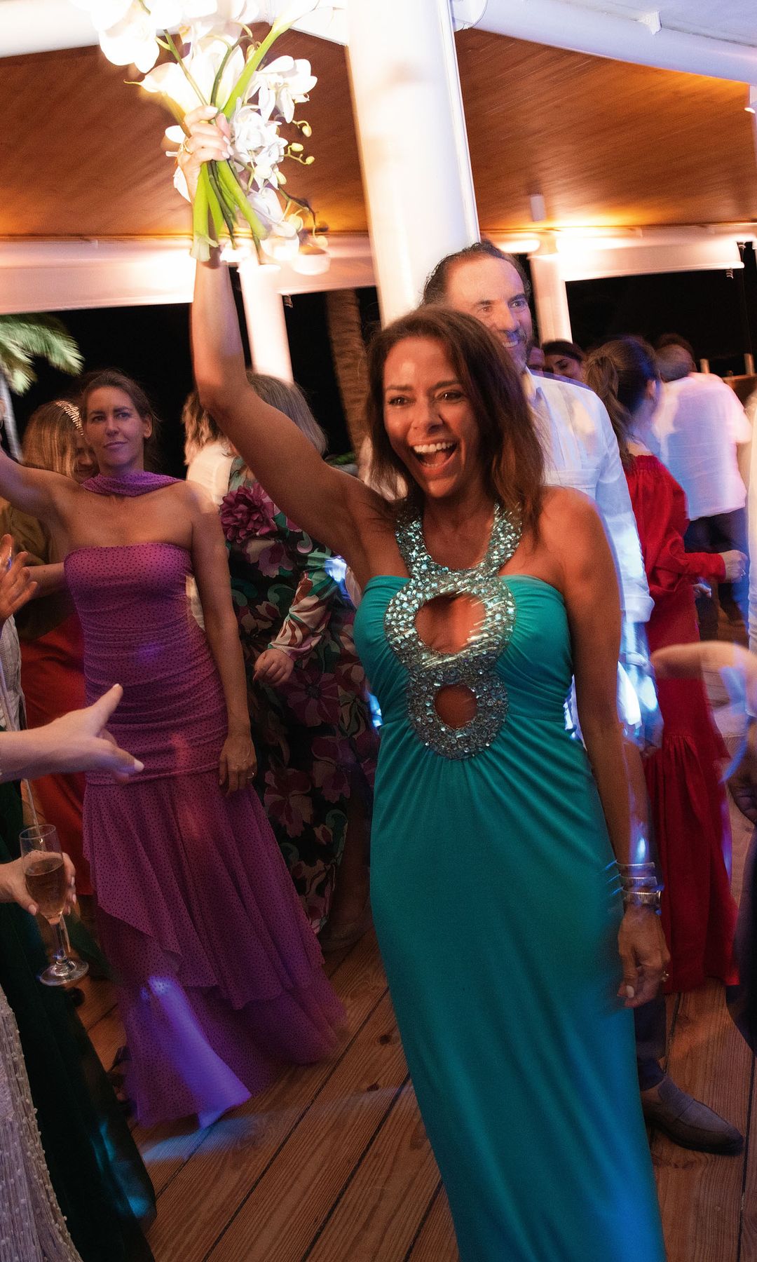 En una fiesta, una mujer con vestido azul sostiene flores. Varios invitados, incluyendo un hombre en camisa blanca, bailan y ríen.