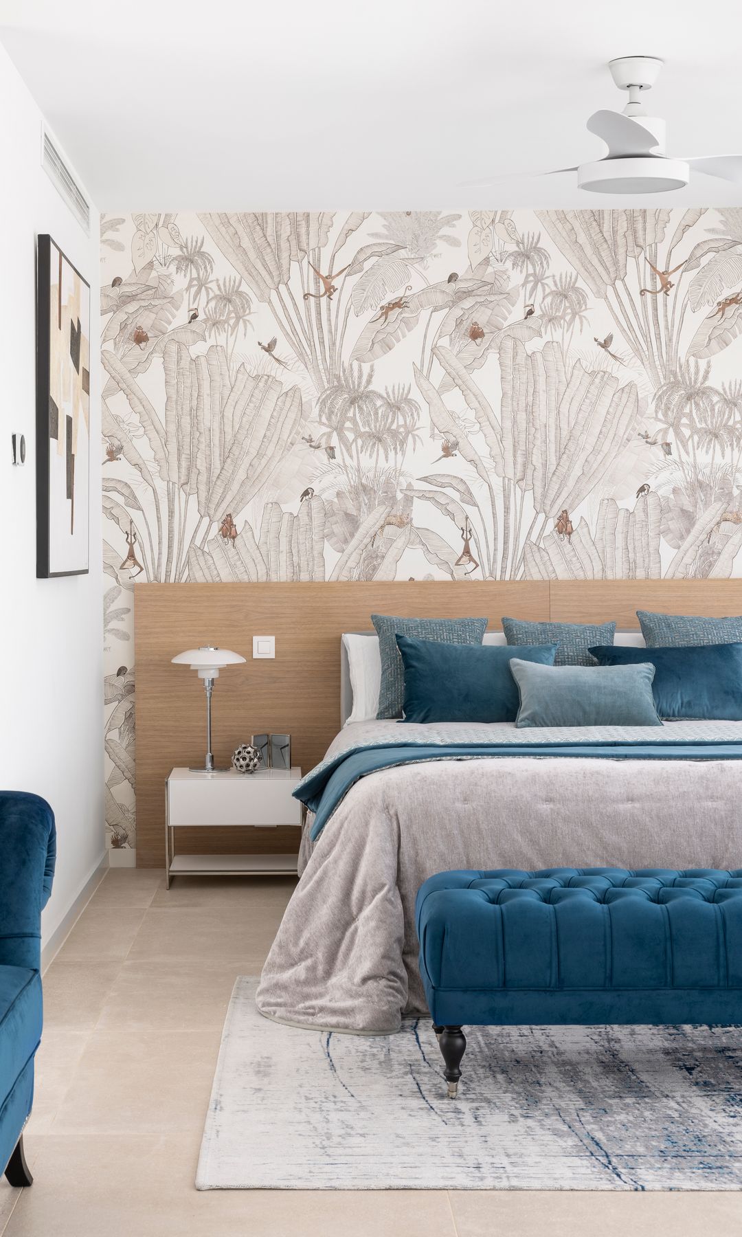 Dormitorio con cabecero de madera, banco a los pies de la cama de capitoné azul, sofá de terciopelo azul, alfombra, cojines azules sobre la cama, papel pintado con monos en tonos marrones