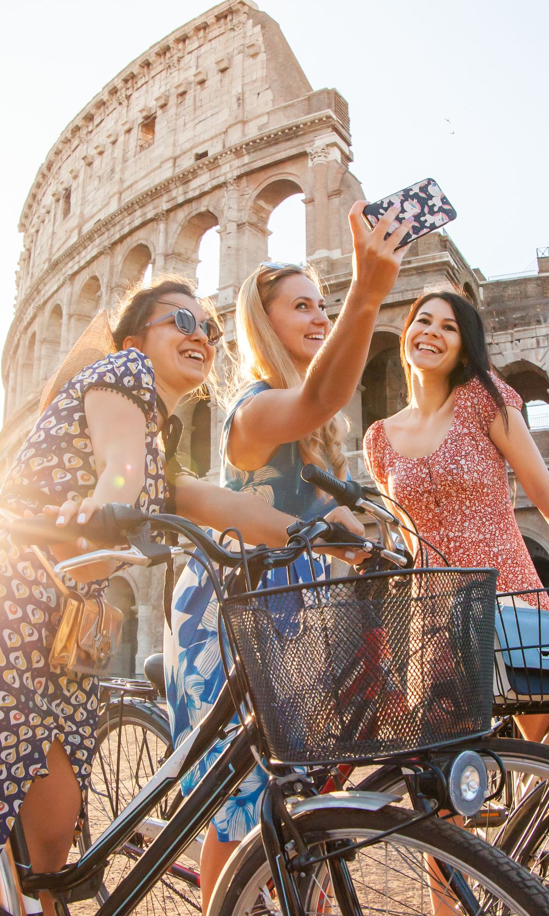 tres chicas sonrientes en bicicleta haciéndose un selfie junto al Coliseo