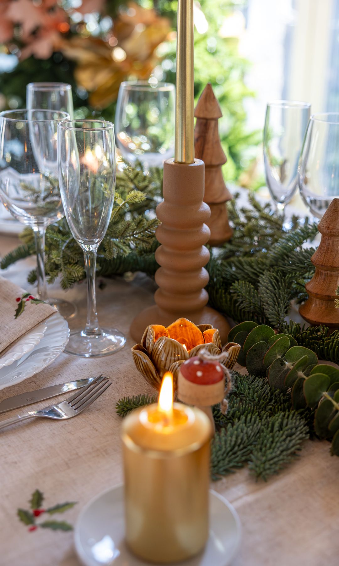 Mesa de Navidad con mantel blanco con motivos navideños, velas doradas, guirnalda verde, copas y candelabros 