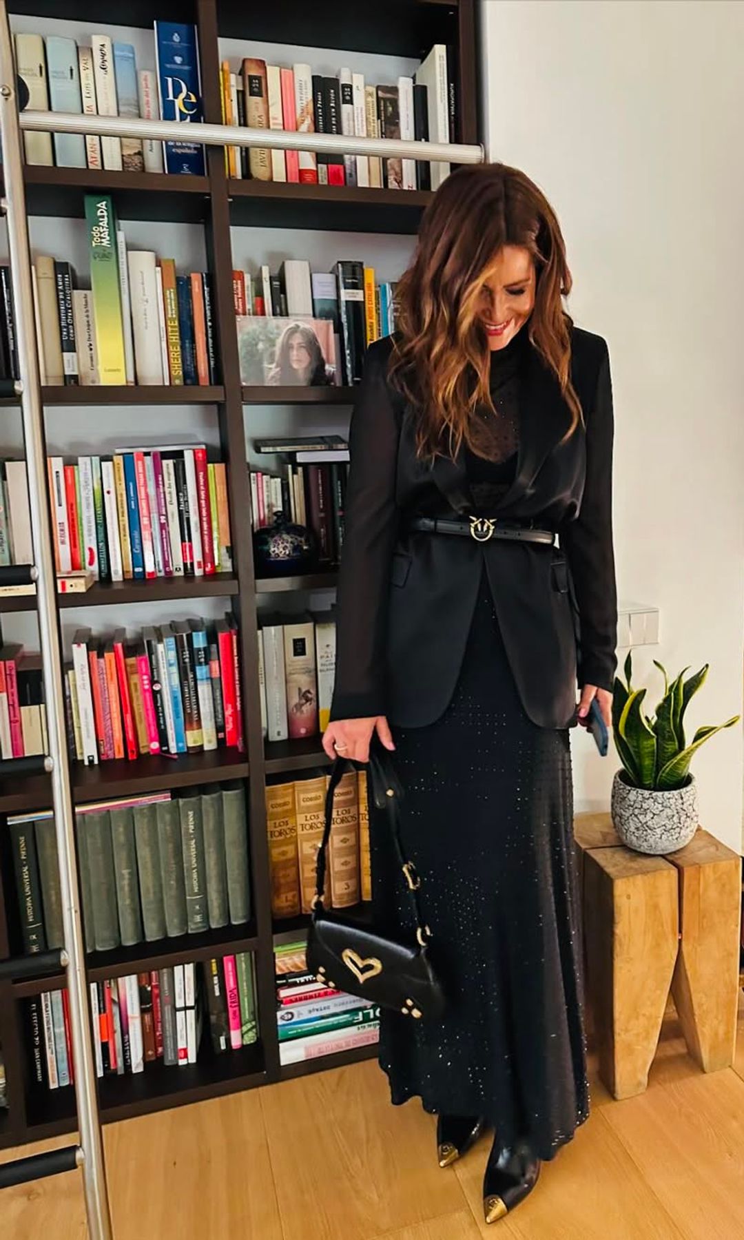 Nuria Roca en un rincón de su casa con estantería negra, libros, plantas, taburete de madera y suelo de madera 