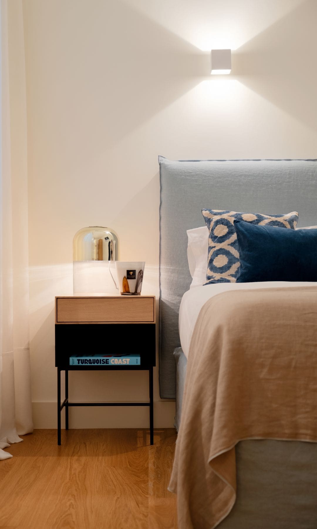 Dormitorio con cabecero tapizado gris, colcha color tierra a los pies, cojines de color azul, lisos y con motivos geométricos, aplique de pared, mesita de noche