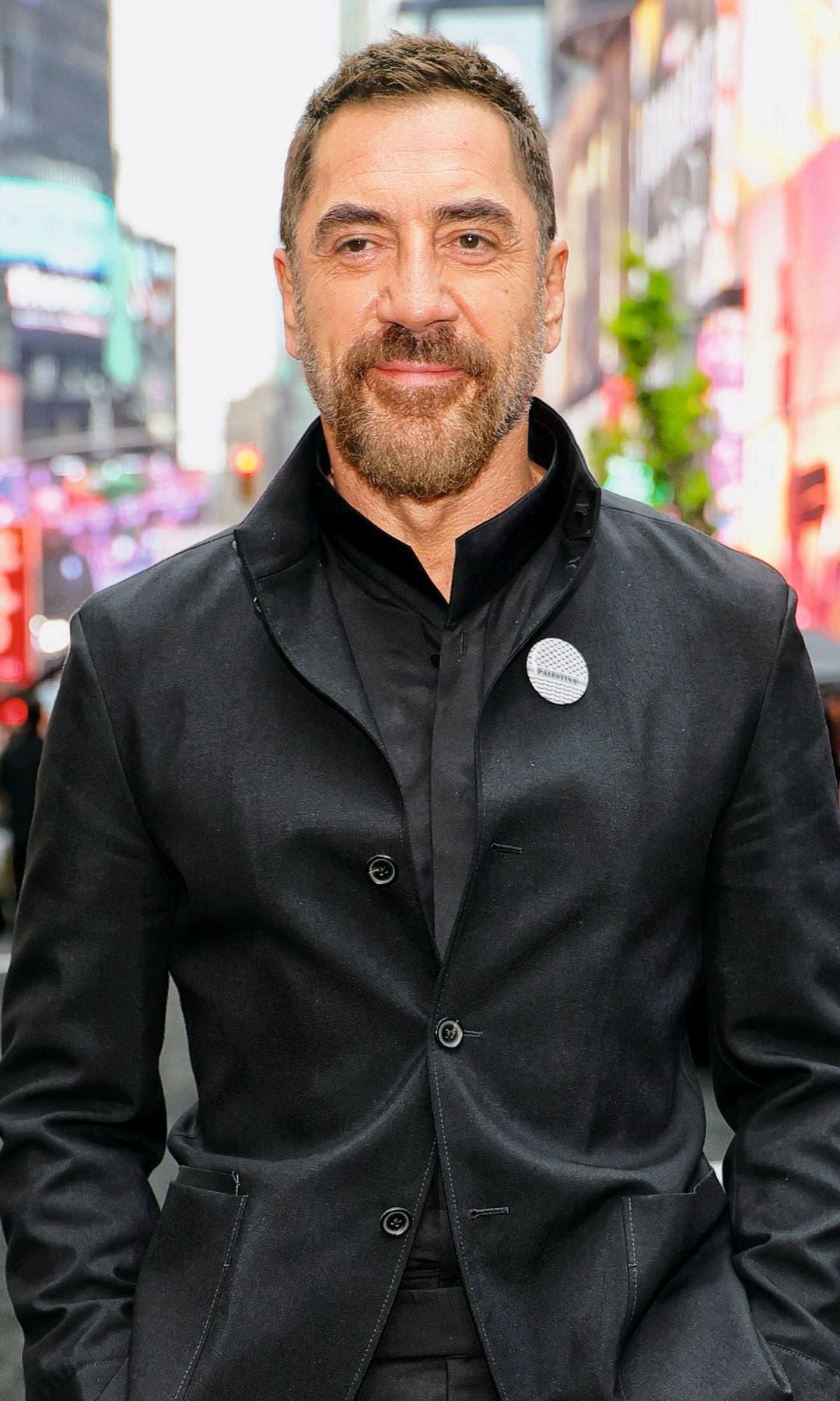 Javier Bardem en traje negro, sonriendo en una calle urbana con coches de carreras.