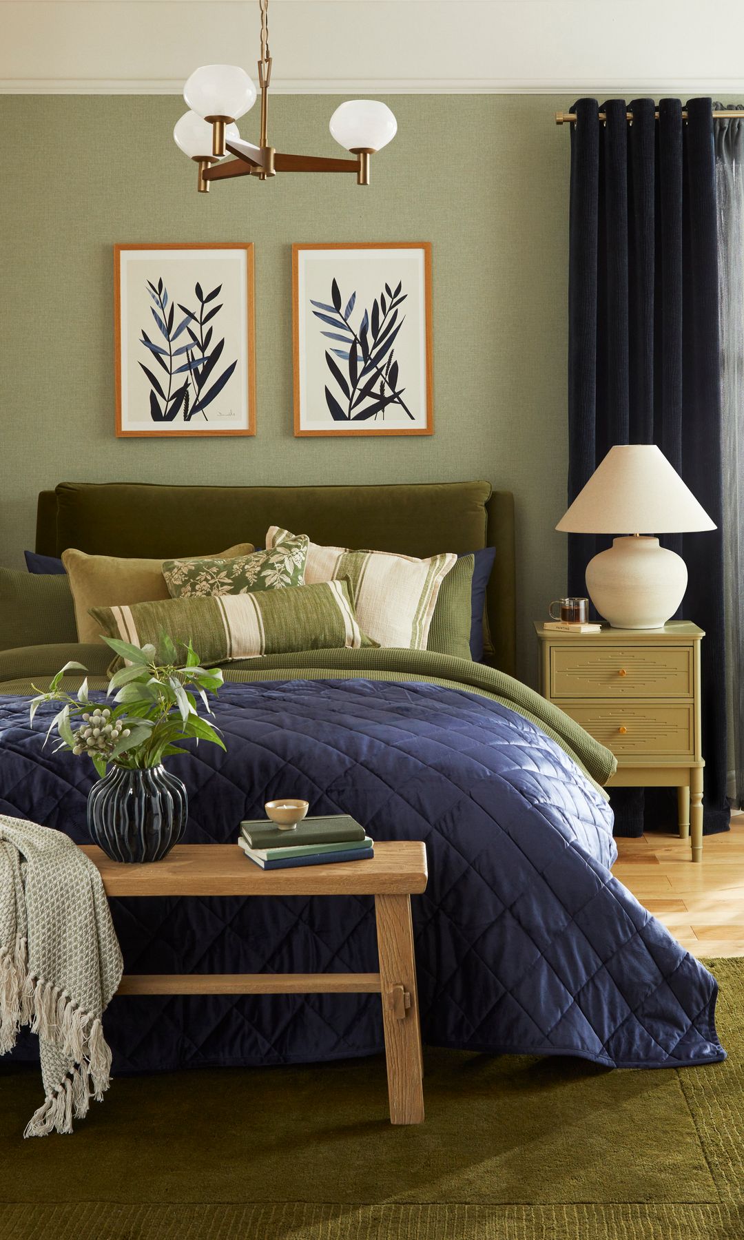 Dormitorio con paredes verdes, ropa de cama verde, funda nórdica azul, mesita de noche, lámpara de sobremesa, banco de madera a los pies de la cama 