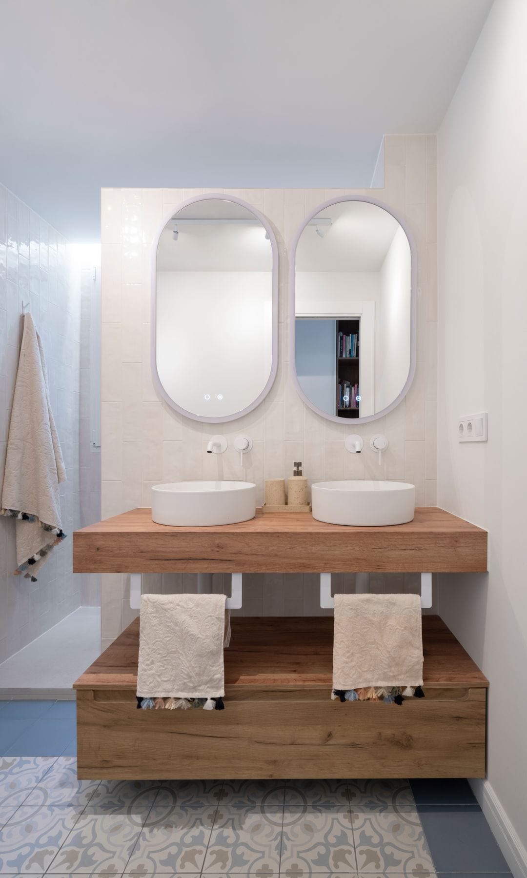 Baño compartido con dos lavabos, dos espejos, mueble bajolavabo abierto en la parte central, dos toalleros blancos con toallas con borlas, suelo de baldosas a modo hidráulico