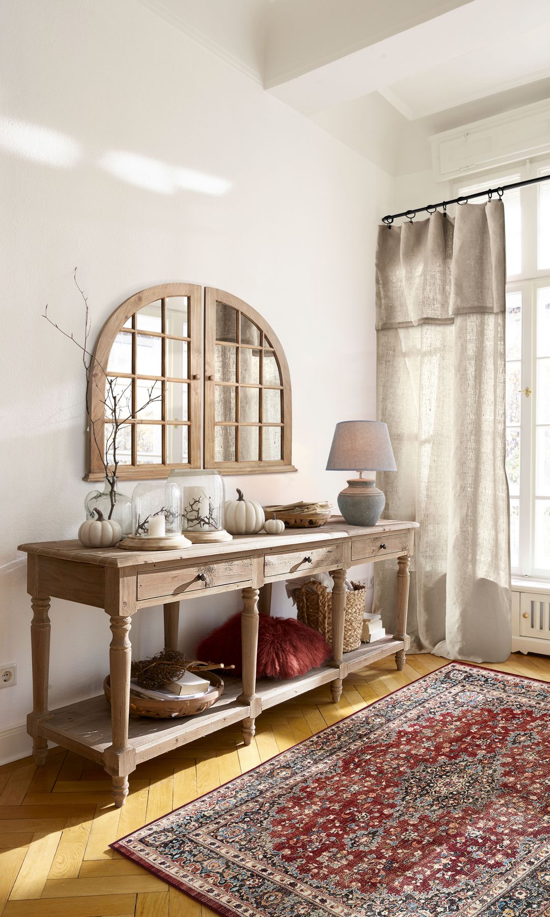 Recibidor de estilo rústico con alfombra, aparador con cajones y parte abierta, cortinas, lámpara de sobremesa, adornos, calabazas