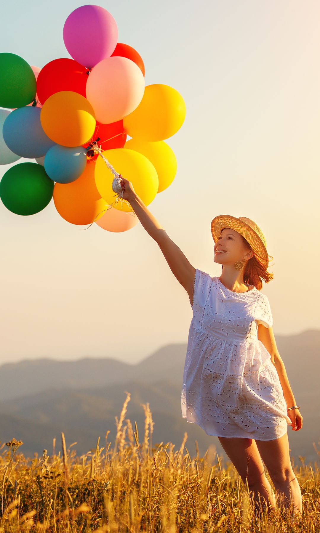 Mujer en vestido blanco y sombrero sosteniendo globos en un campo al atardecer.