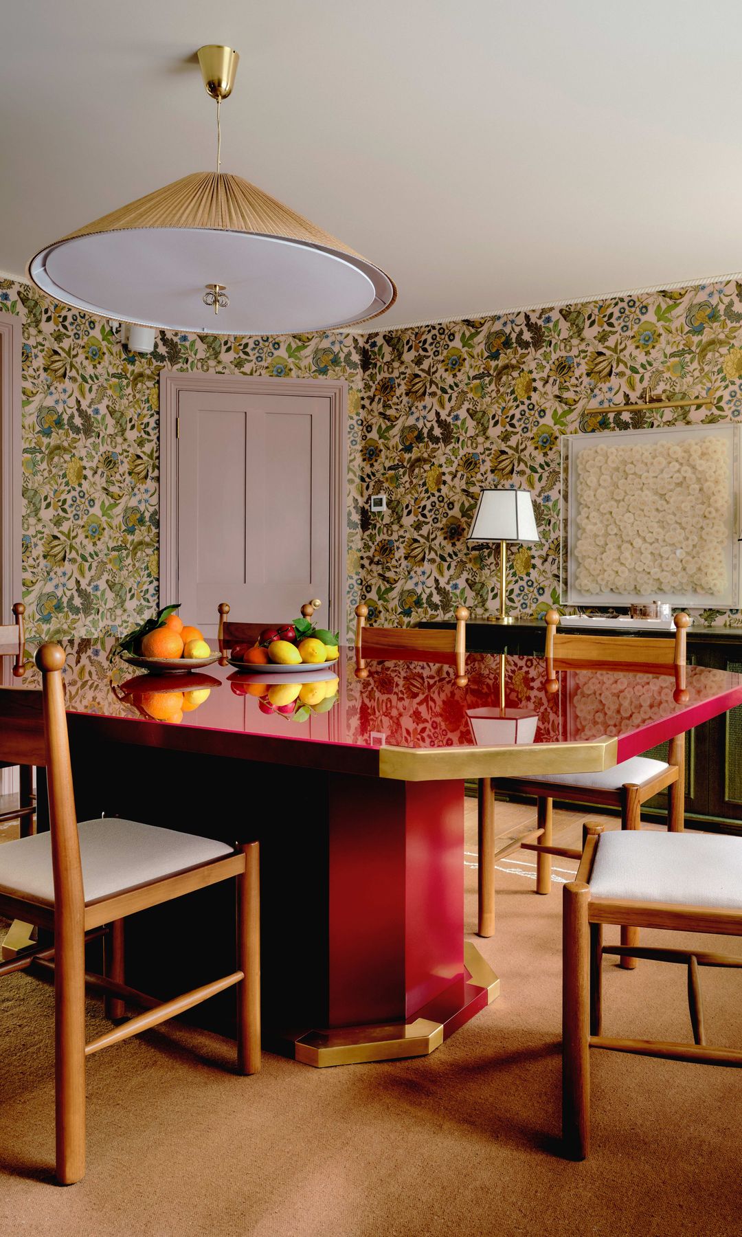 Comedor con papel pintado de flores, mesa de comedor roja, lámpara de techo dorada, frutero en la mesa, sillas de madera con asiento tapizado en blanco, alfombra, aparador verde, lámparas de sobremesa