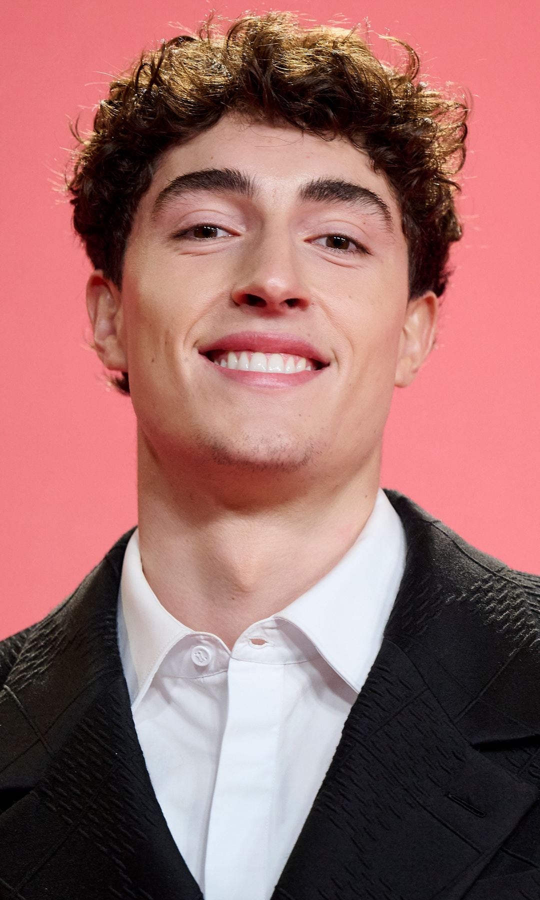  Plex con cabello rizado, camisa blanca y traje negro sonriendo.