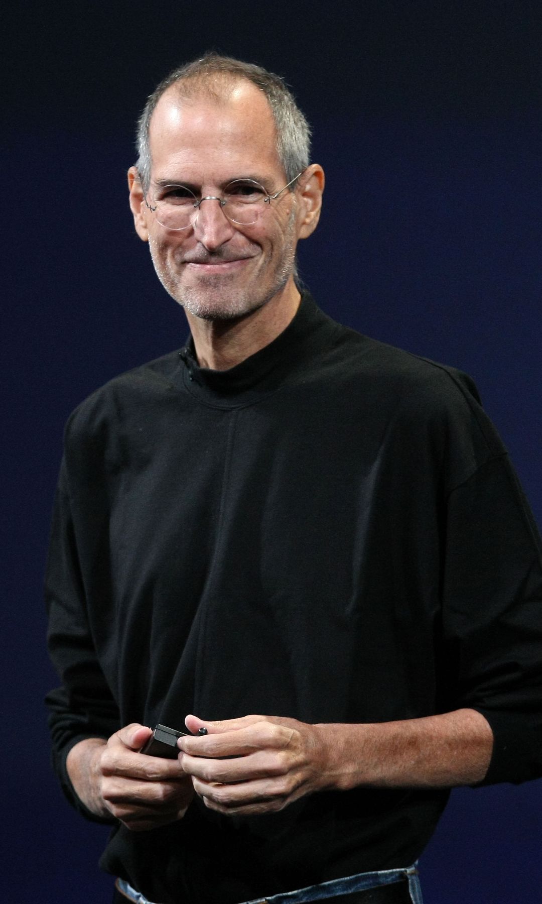Steve Jobs, con camisa negra y gafas, sonríe mientras sostiene un objeto.