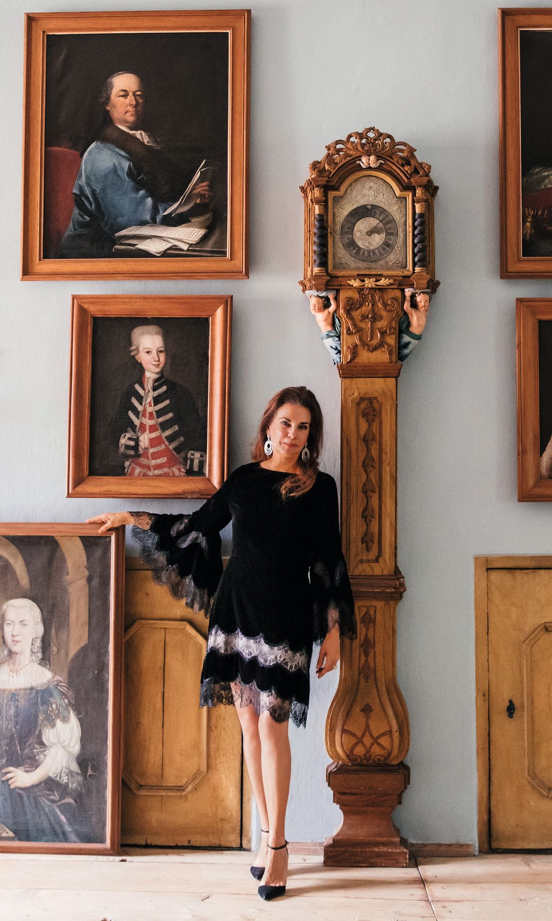 Mujer en vestido negro posa junto a un reloj de pie y retratos en una sala.