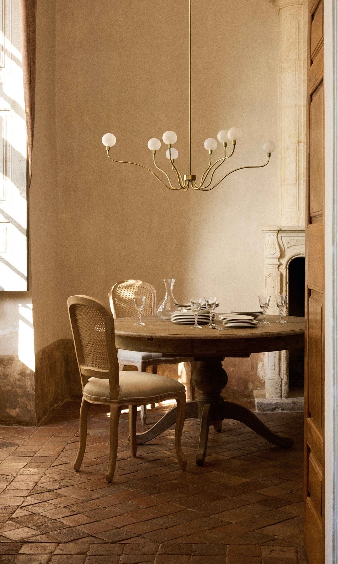 Comedor con chimenea, mesa de comedor redonda, sillas de estilo clásico con rejilla, lámpara de techo, vajilla y copas sobre la mesa, suelo de barro