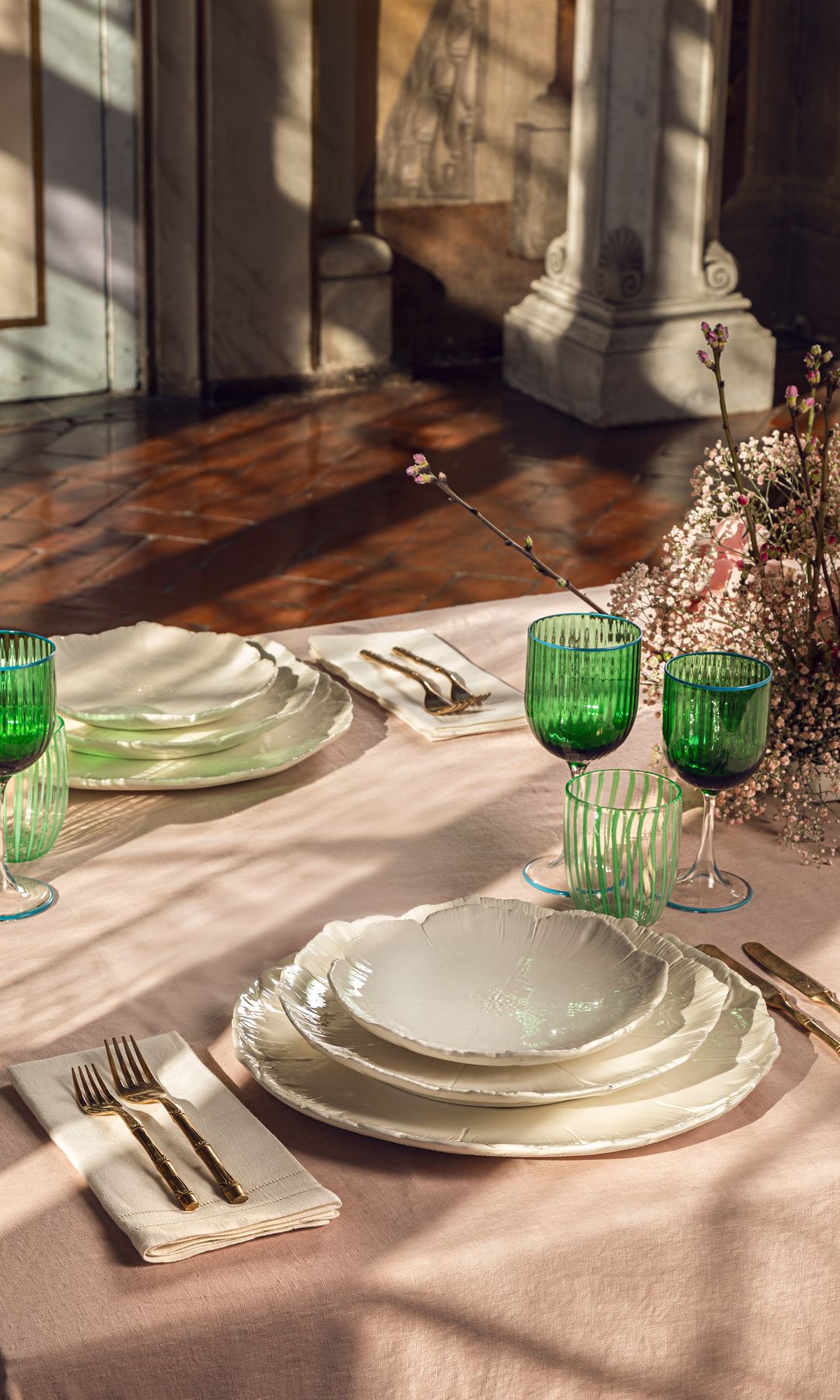 Mesa con mantel rosa claro, cristalería verde, vajilla blanca de porcelana, servilletas blancas, centro de mesa en tonos rosas 