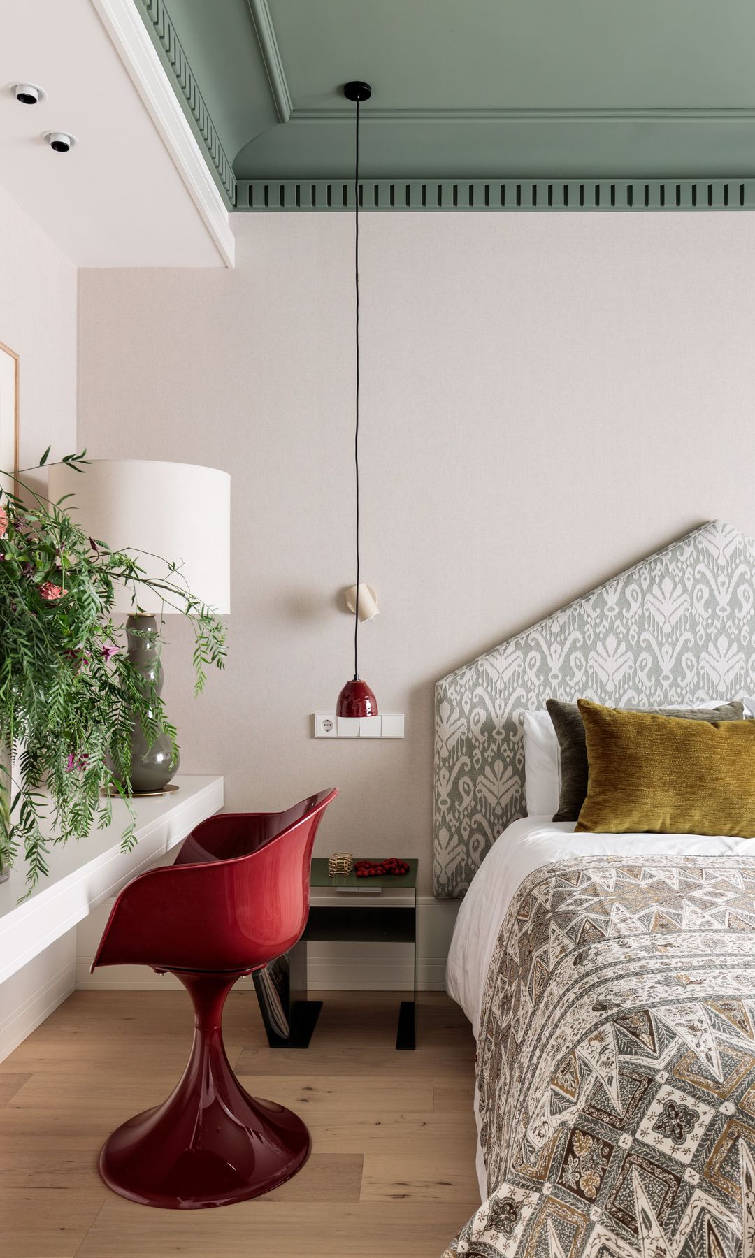 Dormitorio con cabecero tapizado en forma de pico, rincón de tocador con silla roja, lámpara de sobremesa y jarrón con verde