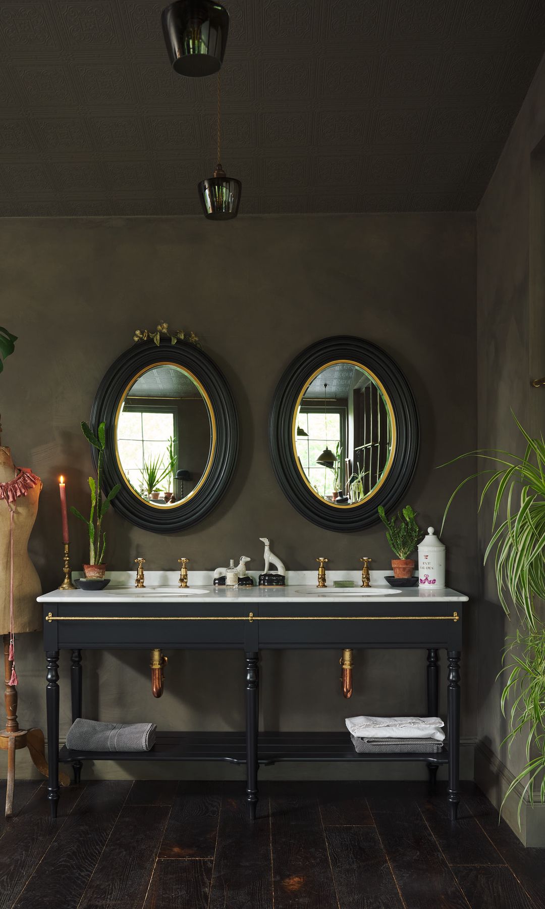 Baño con aires vintage, con espejos ovalados negros y dorados, mueble bajolavabo abierto de color negro, paredes oscuras, plantas colgantes, grifería dorada 