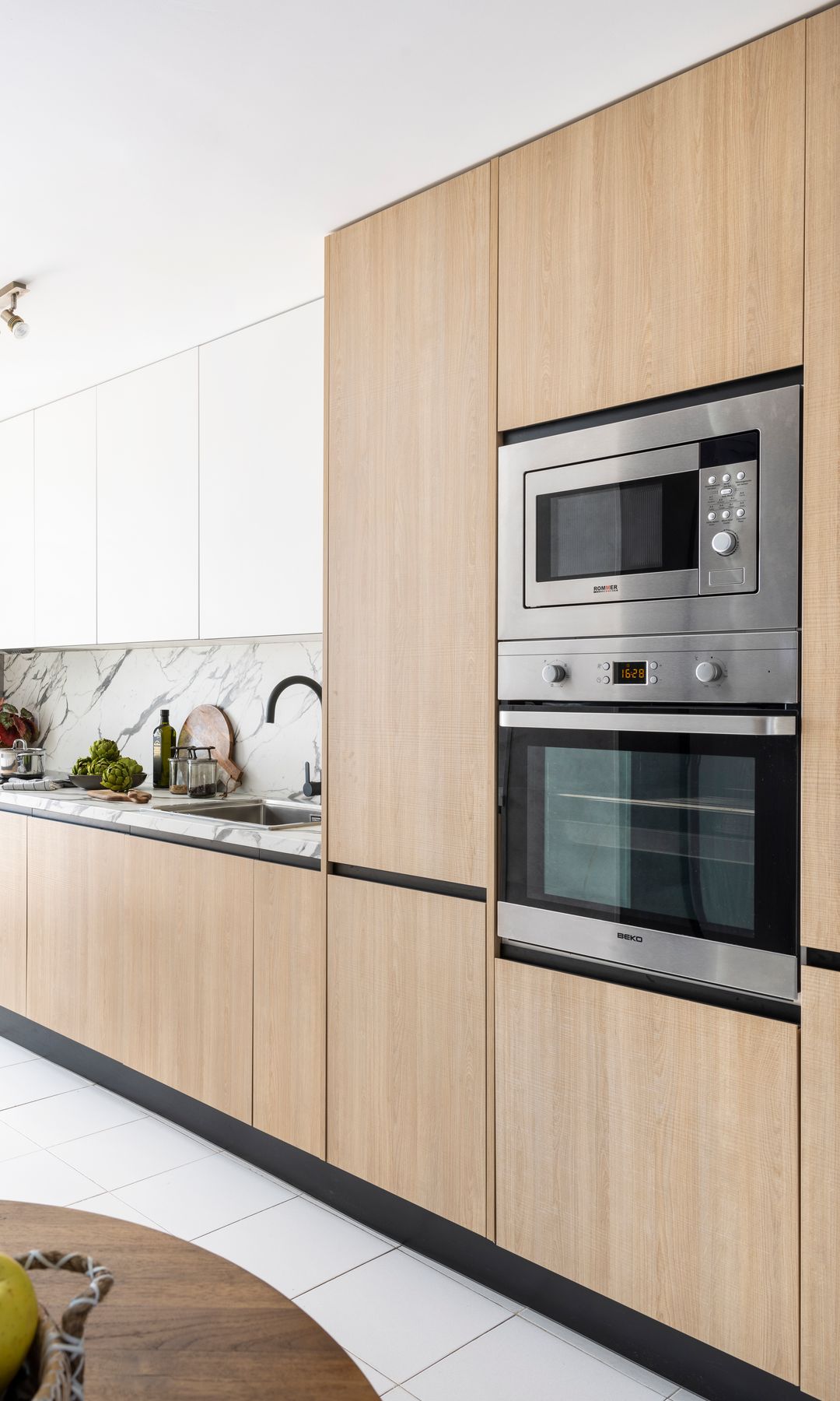 Cocina con muebles de madera y blancos, horno y microondas de acero en columna, suelo blanco, antepecho de azulejos, regleta de focos