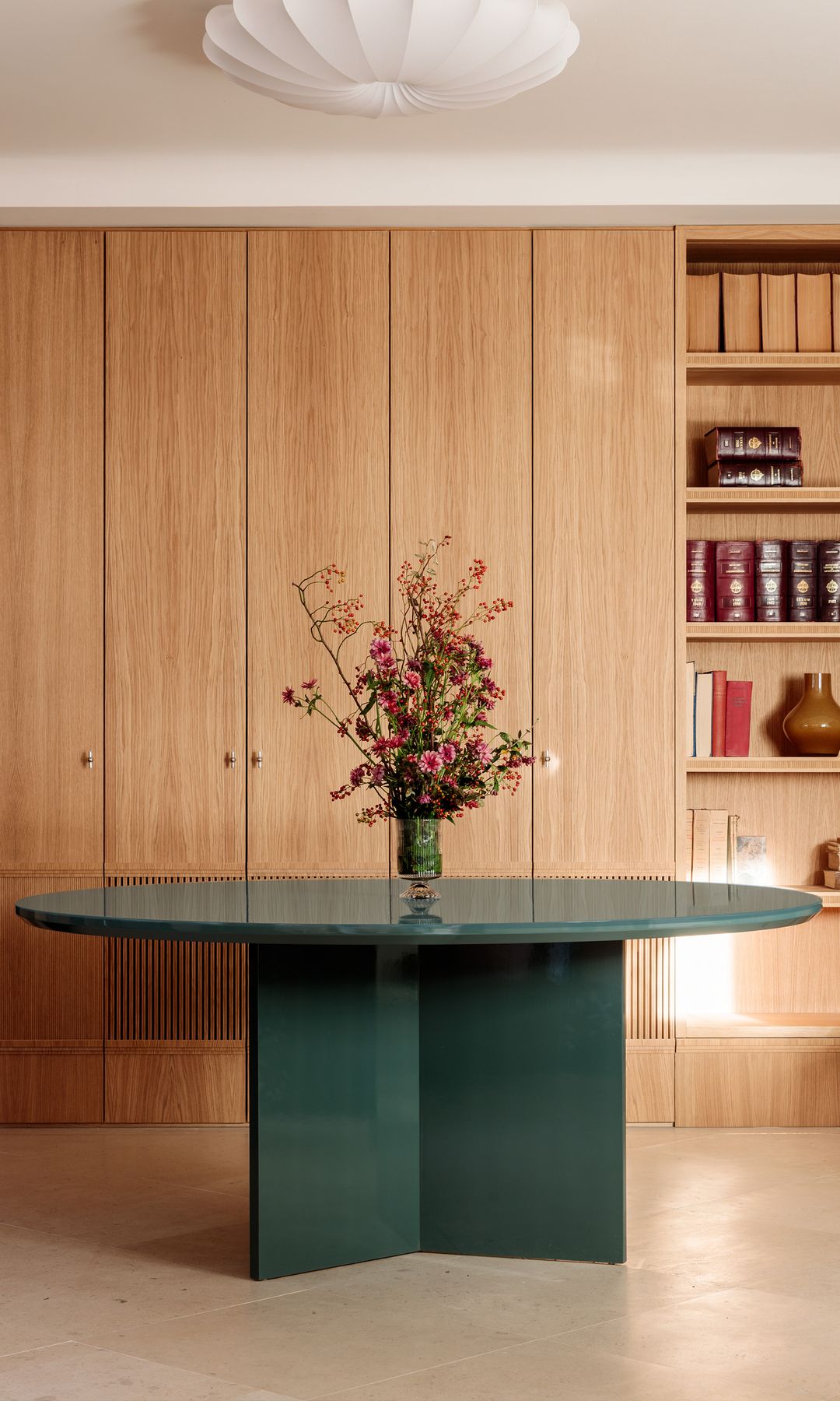 Comedor con mueble de madera a medida con parte con puertas y parte abierta con baldas, mesa de comedor verde con jarrón con flores, lámpara de techo