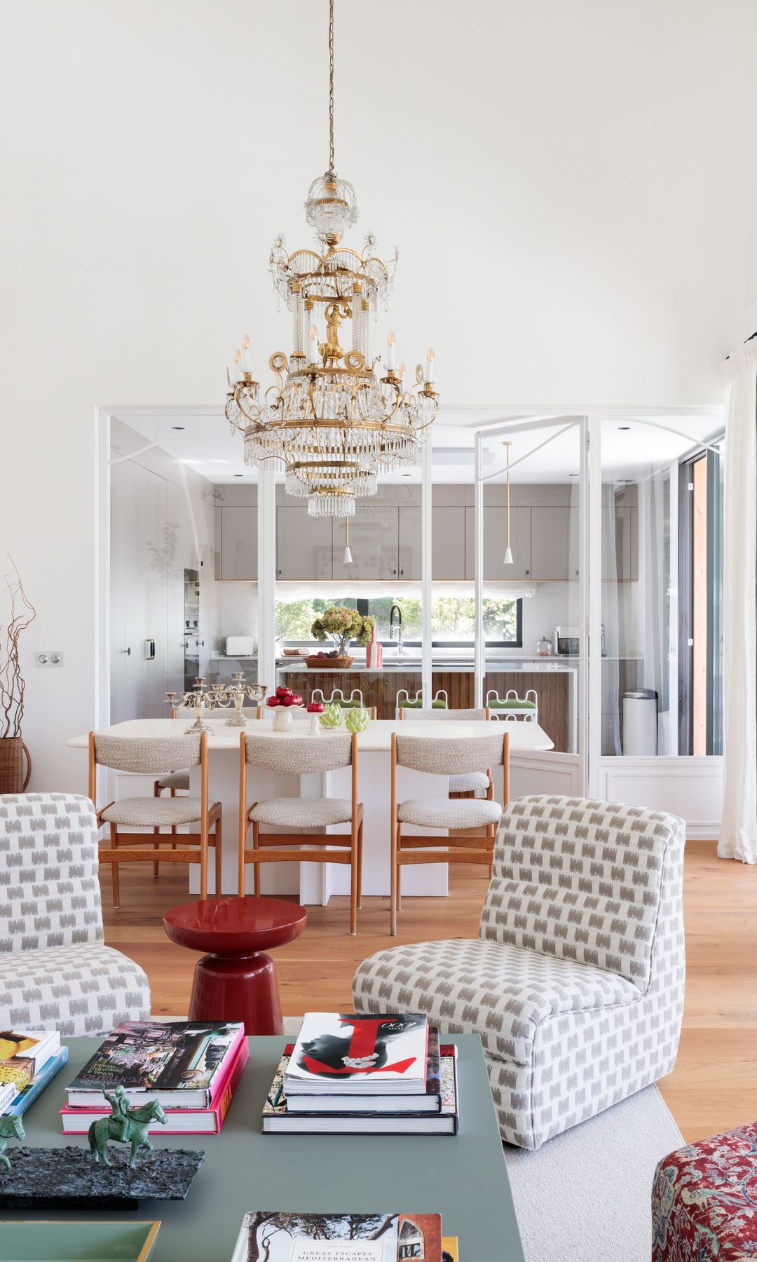 Salón comedor con lámpara de araña, butaca, mesa de centro gris con revistas y libros, mesa de comedor blanca, sillas de madera tapizadas en blanca, suelo de madera, taburete rojo