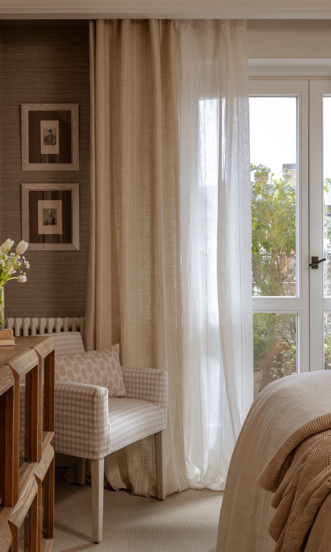 Dormitorio con aparador con vitrina baja de madera, tipo aparador, jarrón con flores, cortinas y visillos blancos, butaca tapizada, cuadros en la pared
