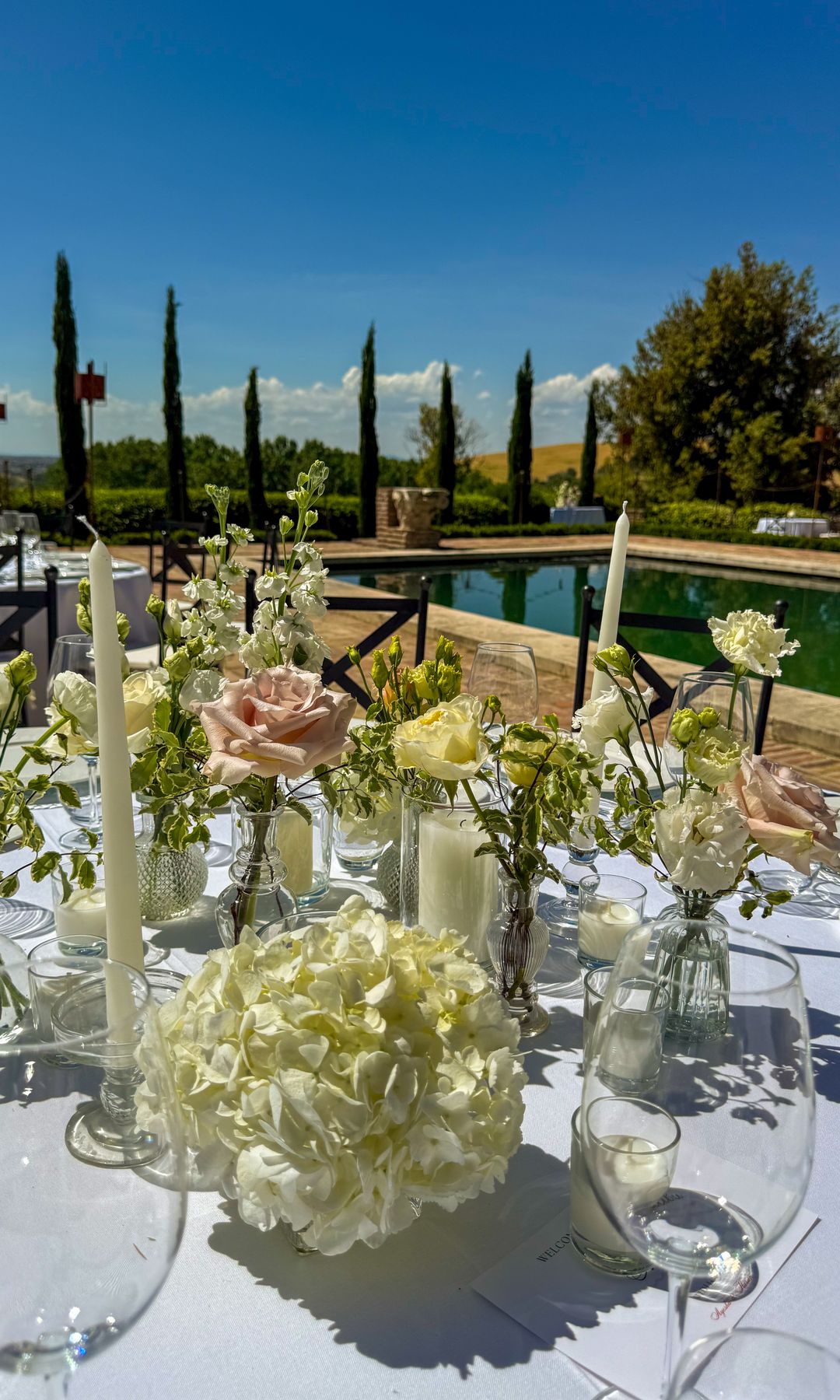 Mesa al aire libre junto a la piscina con mantel blanco, centros de mesa, flores y copas