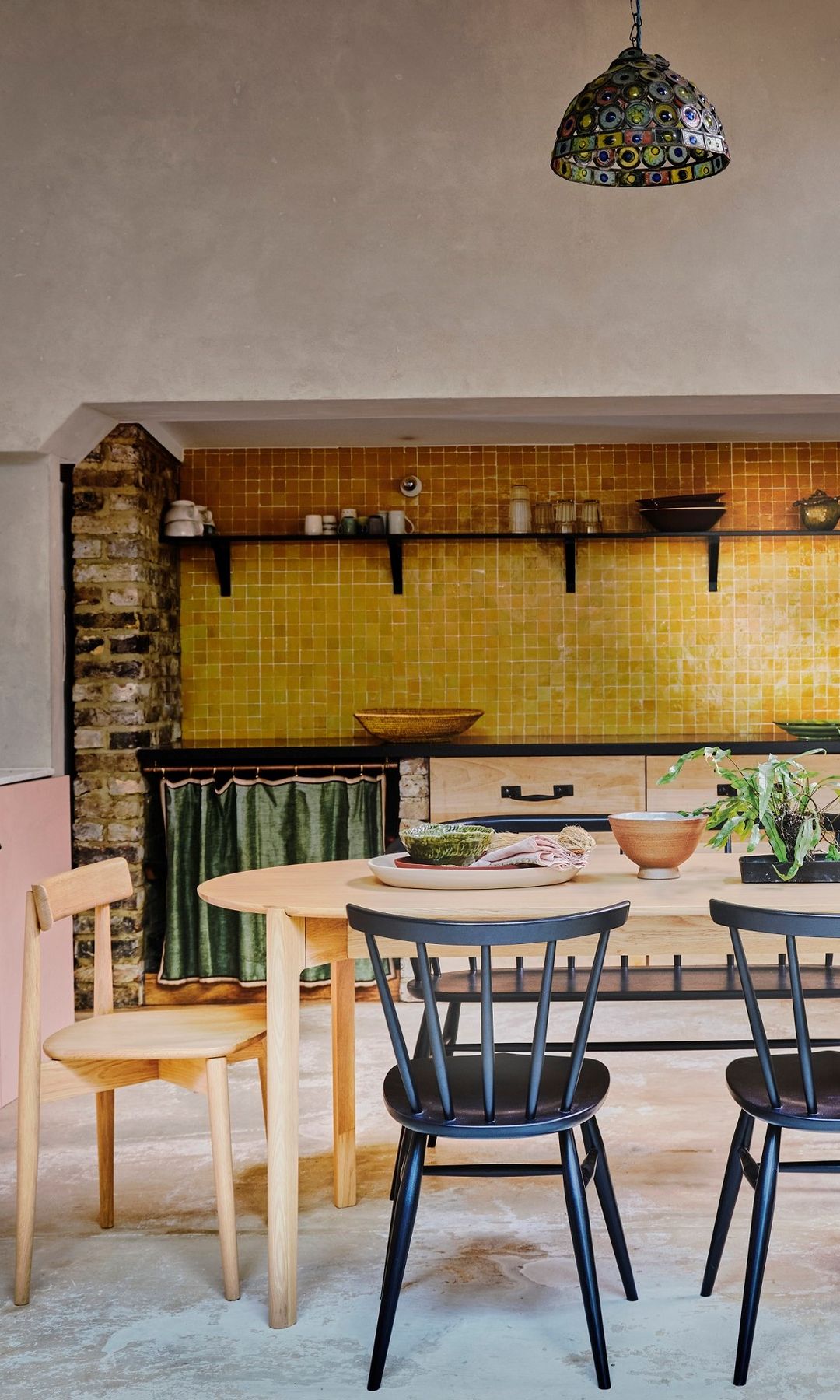 Cocina sin armarios superiores, con muebles rosas, mesa de madera con sillas de comedor negras con barrotes de madera y de madera natural, platos y fuentes