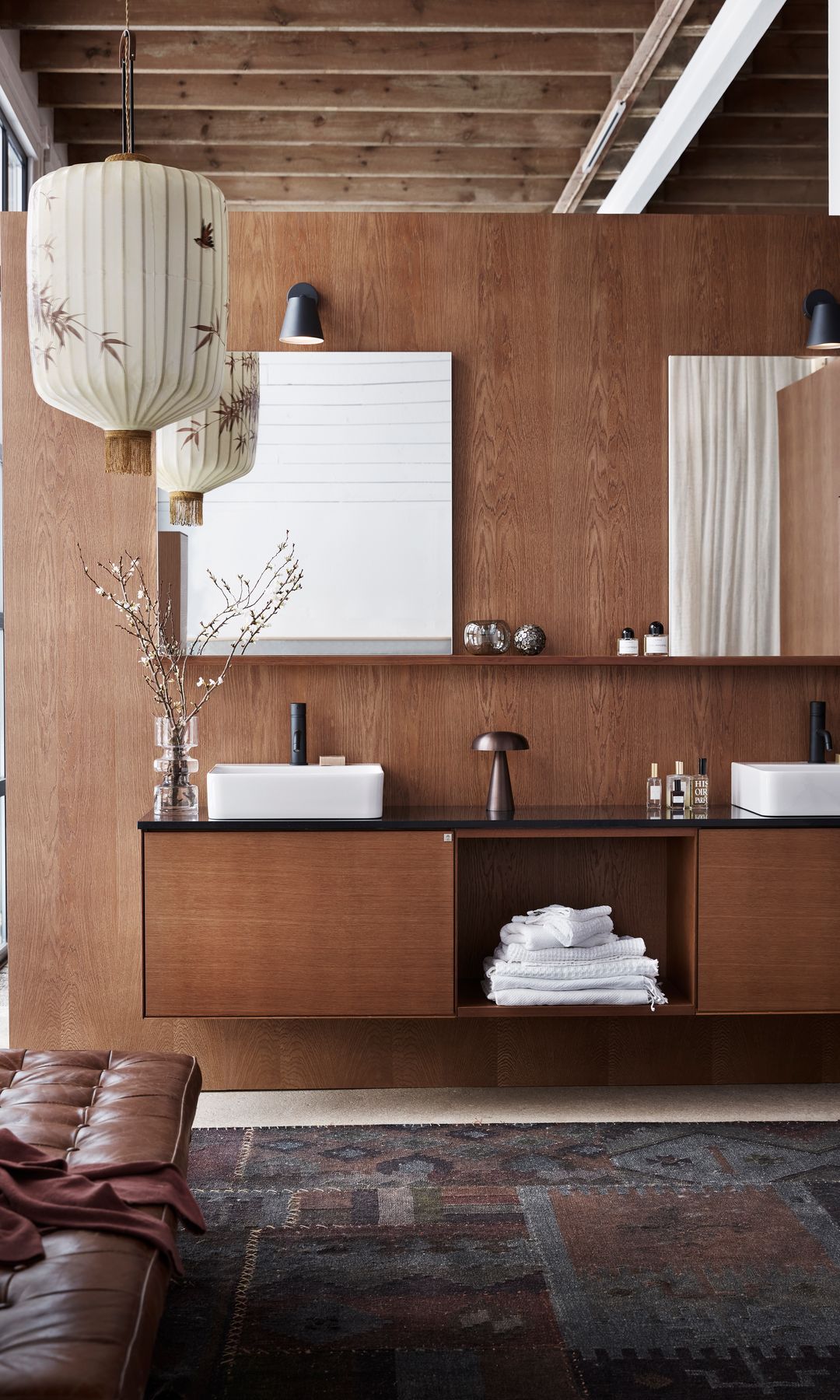 Baño de estilo japandi, con muebles de madera oscura, espejos, doble lavamanos, banco de cuero, alfombra, lámpara de papel, tipo farolillo