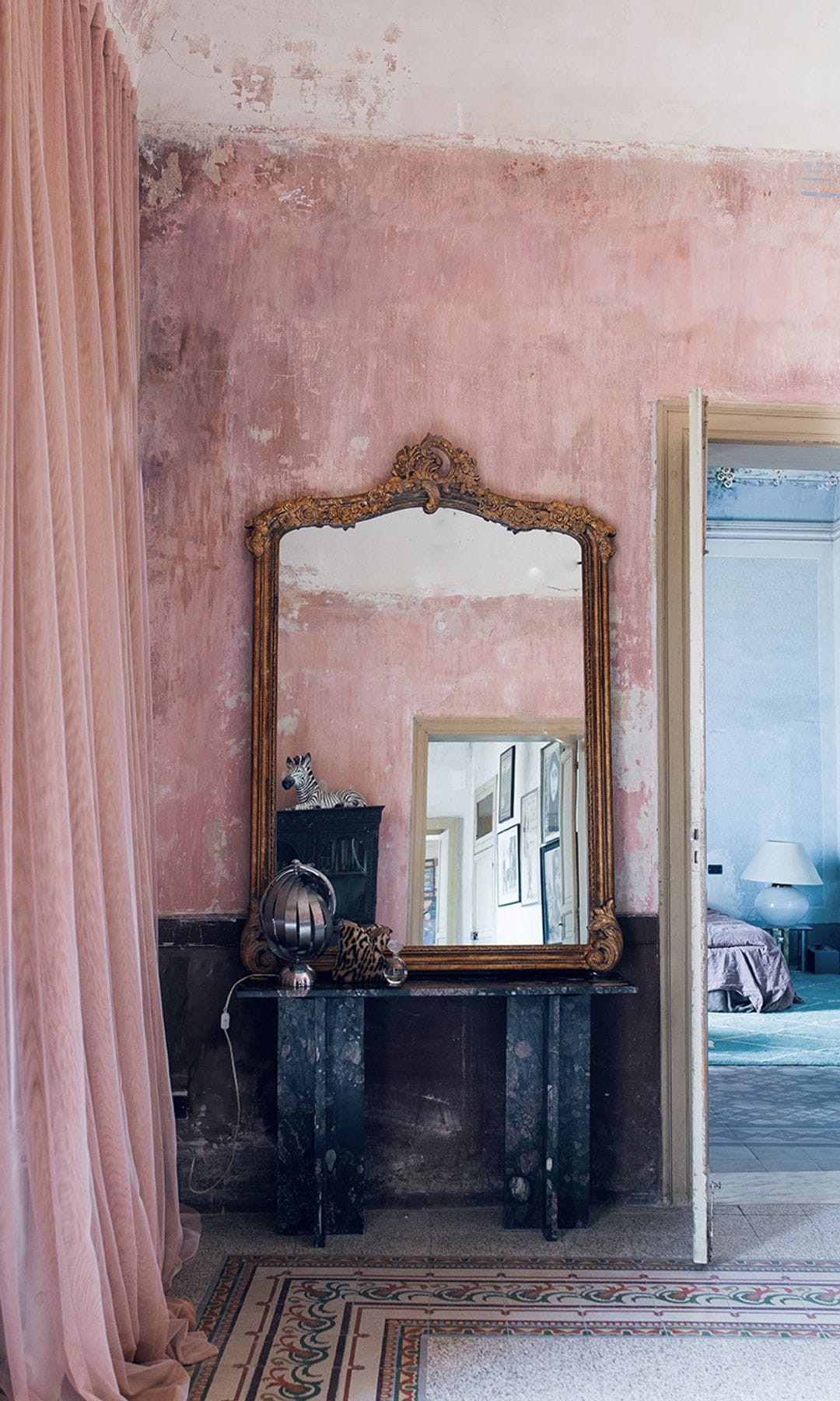 Salón con paredes rosas, cortinas rosas, suelo hidráulico, espejo dorado antiguo, mesa negra, lámpara de sobremesa metálica