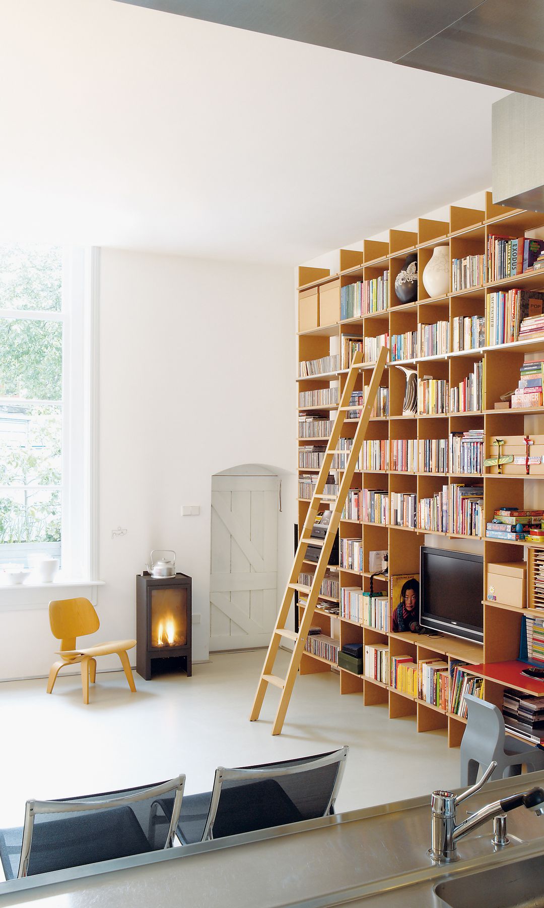 Salón con librería de suelo a techo de madera con escalera abierto a la cocina de acero inoxidable, chimenea exenta y silla de madera 