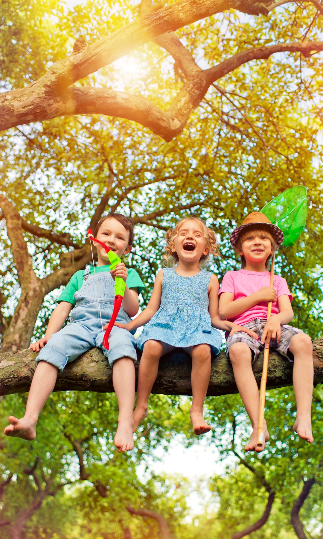 Niños jugando al aire libre subidos a un árbol con un arco de juguete y una red de pesca.
