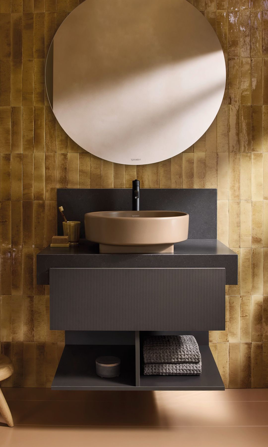 Baño con pared de azulejos, espejo redondo, mueble suspendido con la parte inferior abierta, lavabo redondo sobre la encimera, taburete