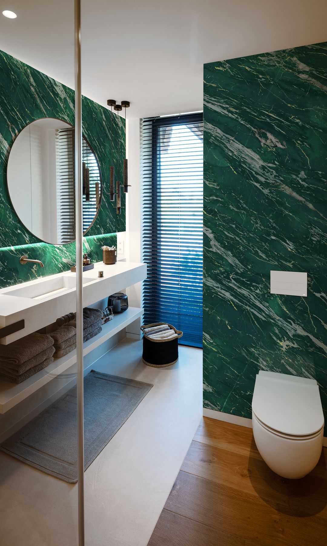 Baño con paredes de mármol verde, suelo de madera, espejo redondo, alfombrilla, ventana, sanitario suspendido 