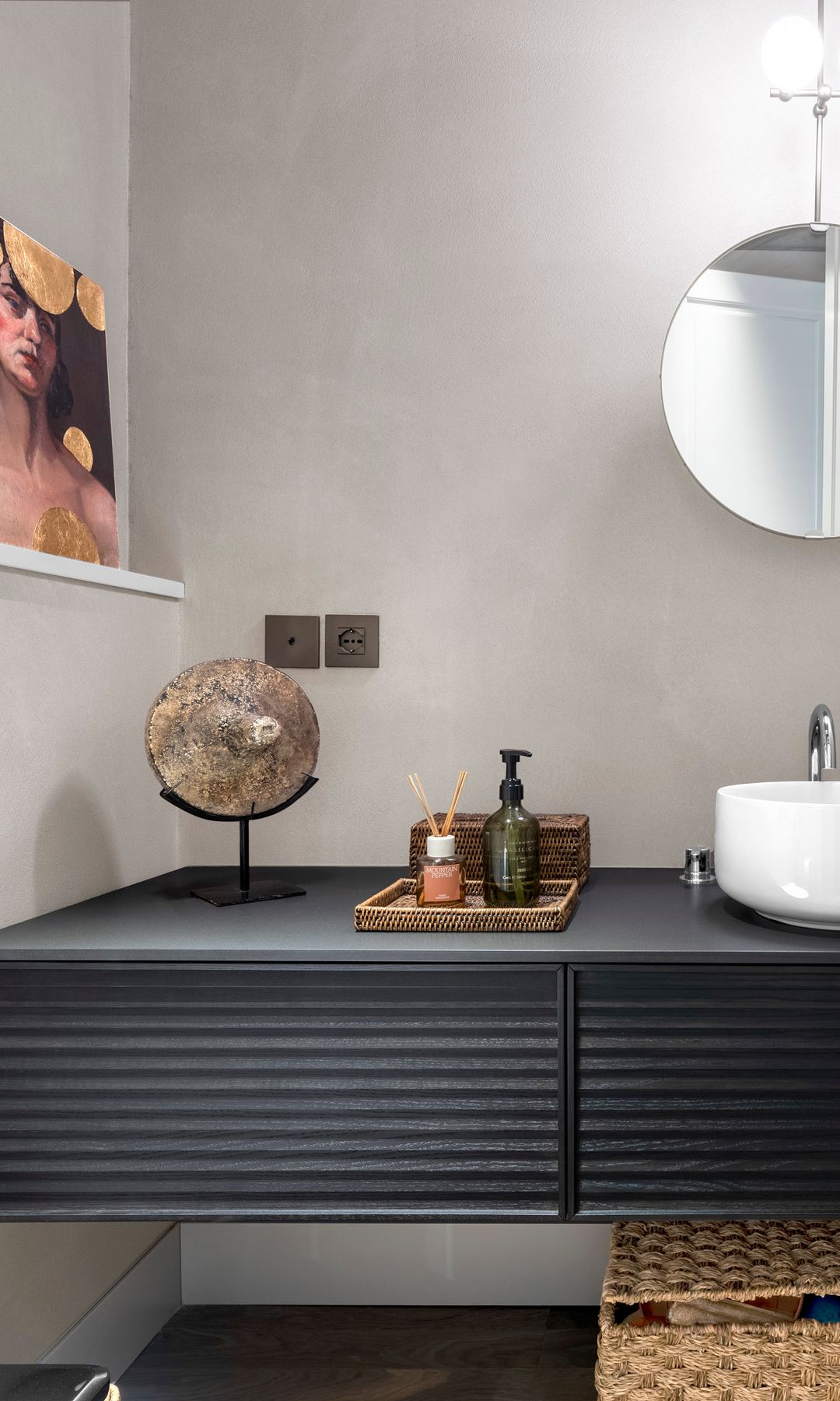 Baño con mueble bajolavabo negro, espejo redondo, lavabo blanco circular, cuadros sobre una repisa