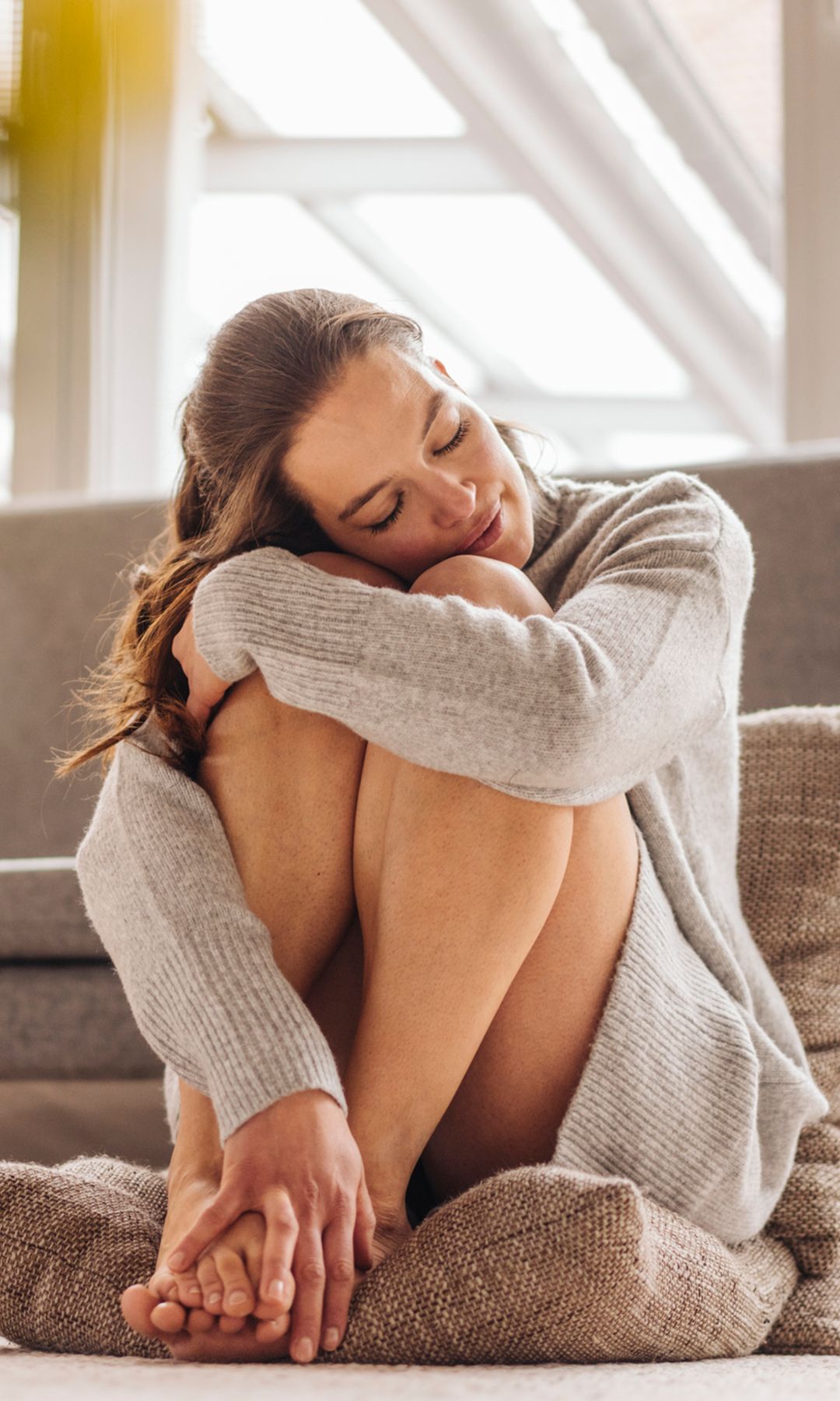 Mujer joven sentada sobre unos cojines en el salón de su casa, con un jersey gris y piernas al aire, abrazándose las rodillas con los ojos cerrados y sonriendo