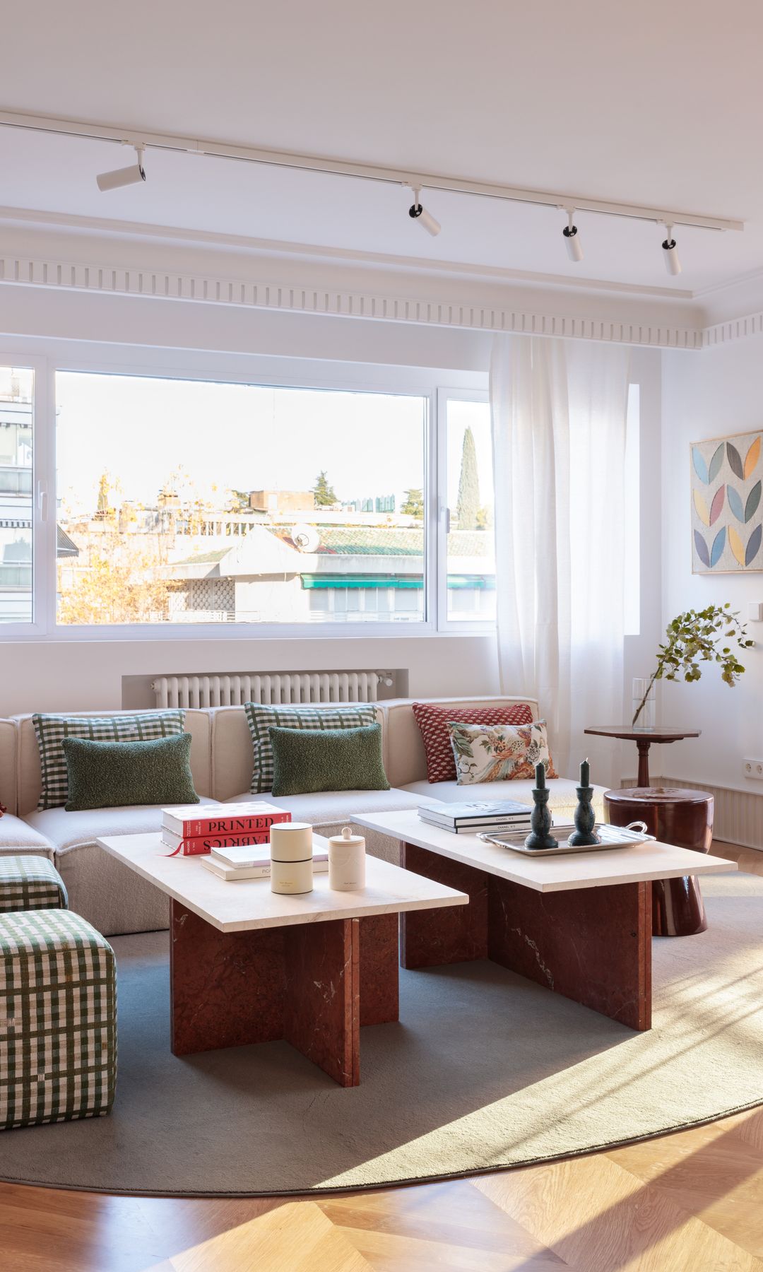Salón con radiador bajo una ventana, visillos blancos, mesa de centro de color rojo con el sobre blanco, cojines sobre el sofá, alfombra y cuadro en la pared