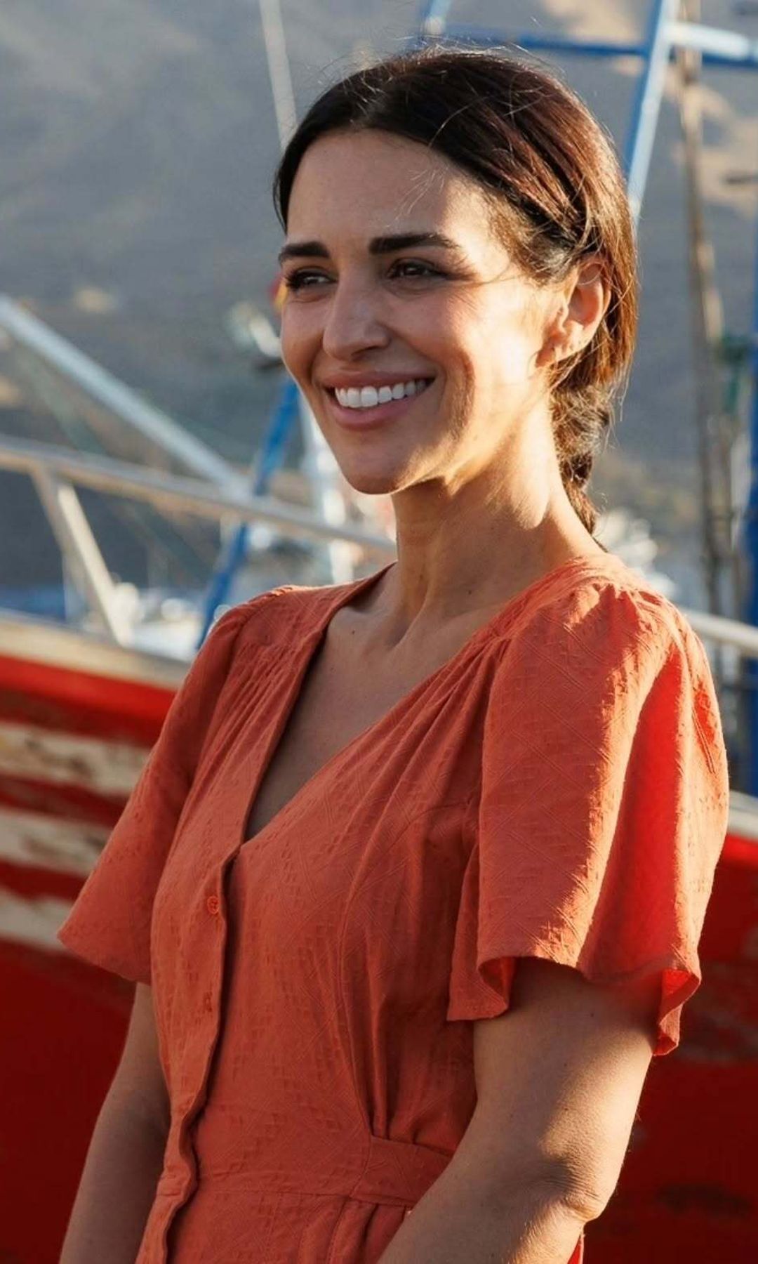 Paula Echevarría sonríe junto a un barco con un vestido naranja y el pelo recogido en una coleta baja