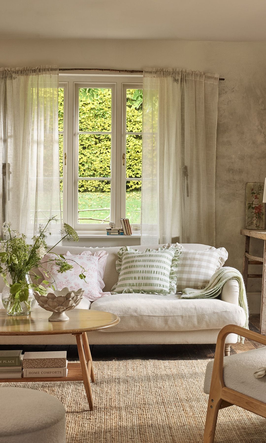 Salón con aire campestre con sofá blanco, cortinas de lino, alfombra de fibras vegetales, cojines de cuadros verdes y beiges y varias plantas 