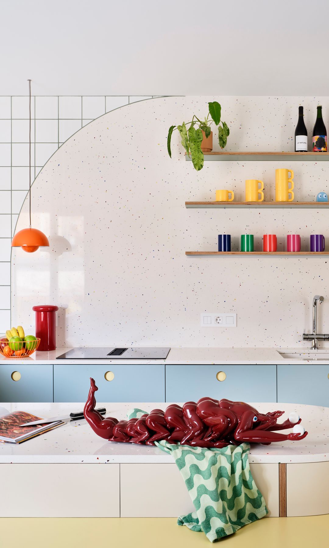 Cocina con muebles azules, baldas de madera con vasos y tazas de colores, lámpara de techo naranja, langosta de cerámica, trapo de cocina verde