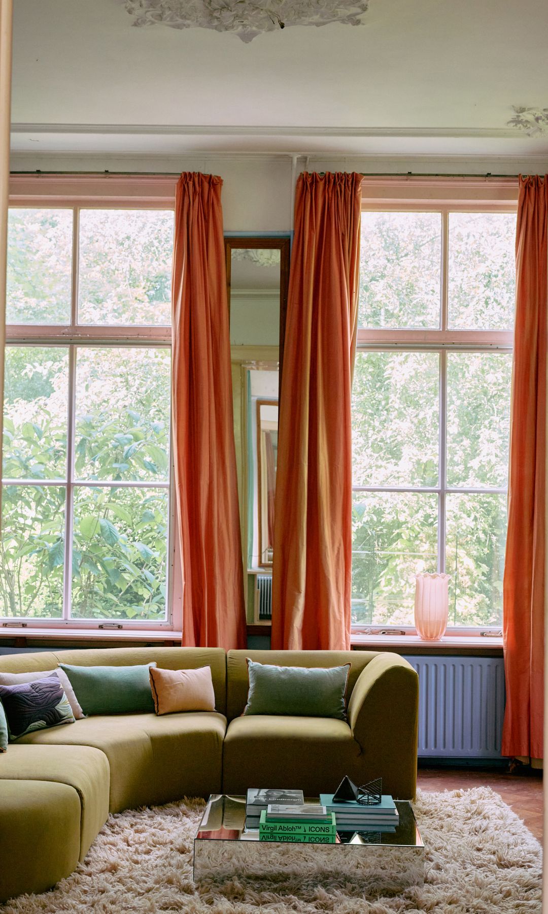 Salón con cortinas naranjas, sofá verde, mesa de centro baja, alfombra de pelo largo blanca, radiador azul, cojines de colores, rosetón en el techo