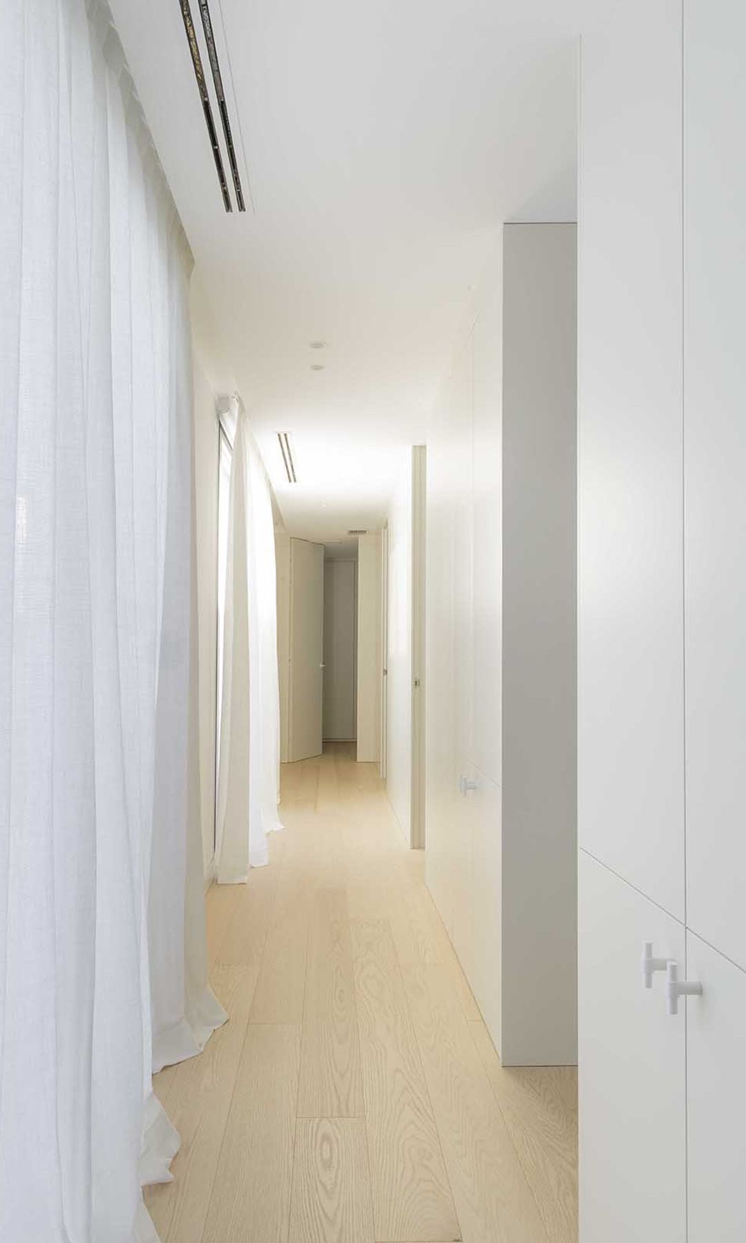Pasillo con armarios blancos en una de las paredes y ventanas con cortinas blancas en el otro, suelo de madera claro 