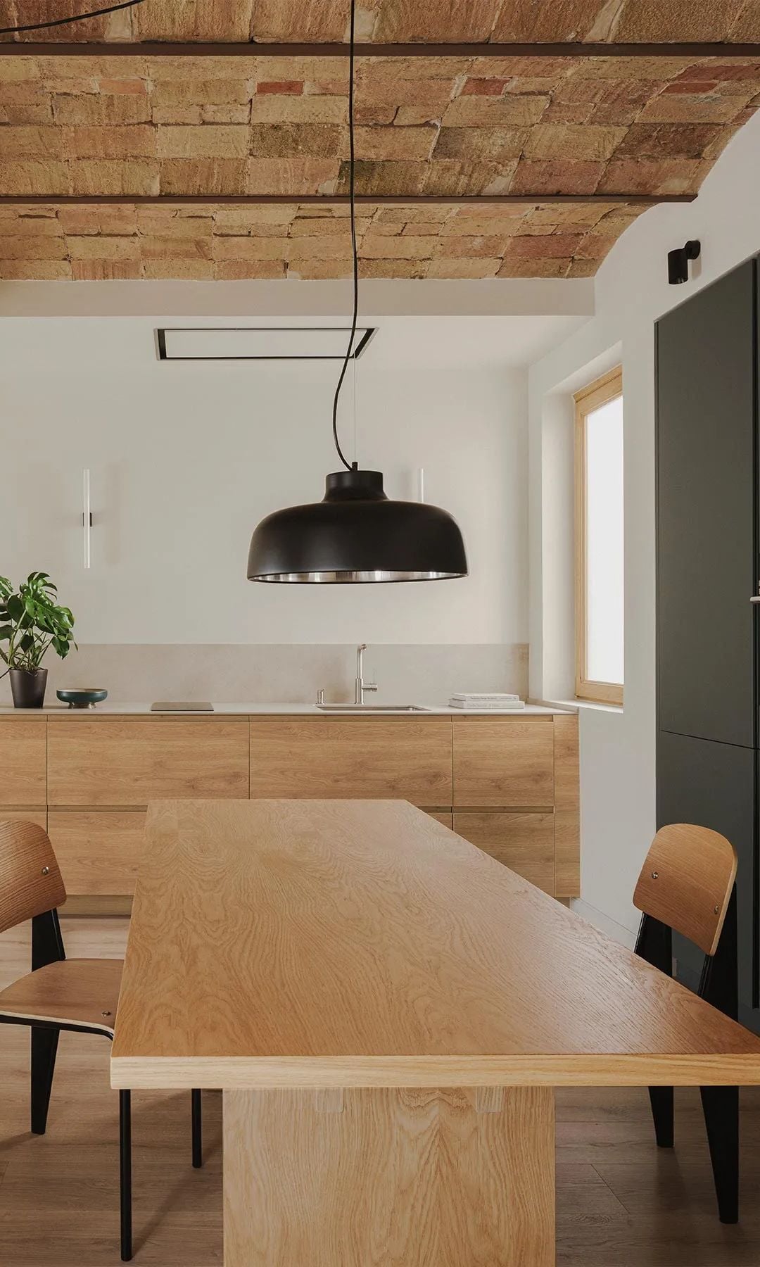 Cocina abierta con comedor, muebles de madera, bóveda catalana en el techo, pared de ladrillo blanca, mesa de madera, lámpara de techo, armarios en gris oscuro