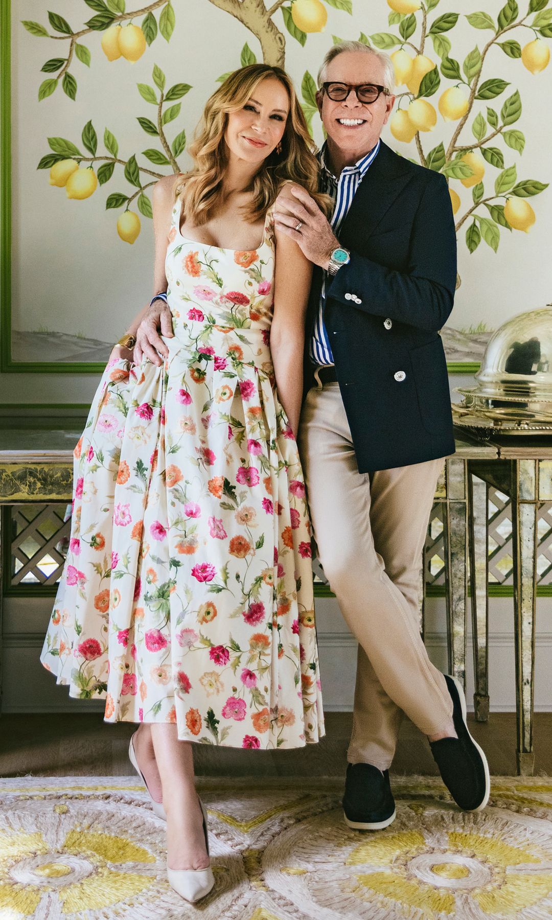 Tommy Hilfiger y su mujer Dee con vestido de flores y traje con fondo de pared con estampado de limoneros y en la esquina una planta encima de un mueble blanco