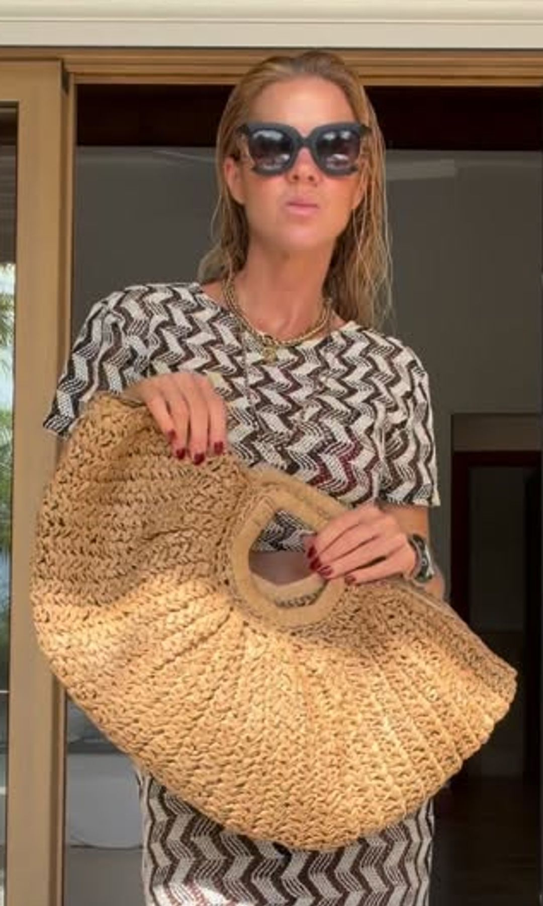 Mujer con cabello largo, gafas de sol y vestido estampado sostiene un bolso tejido.