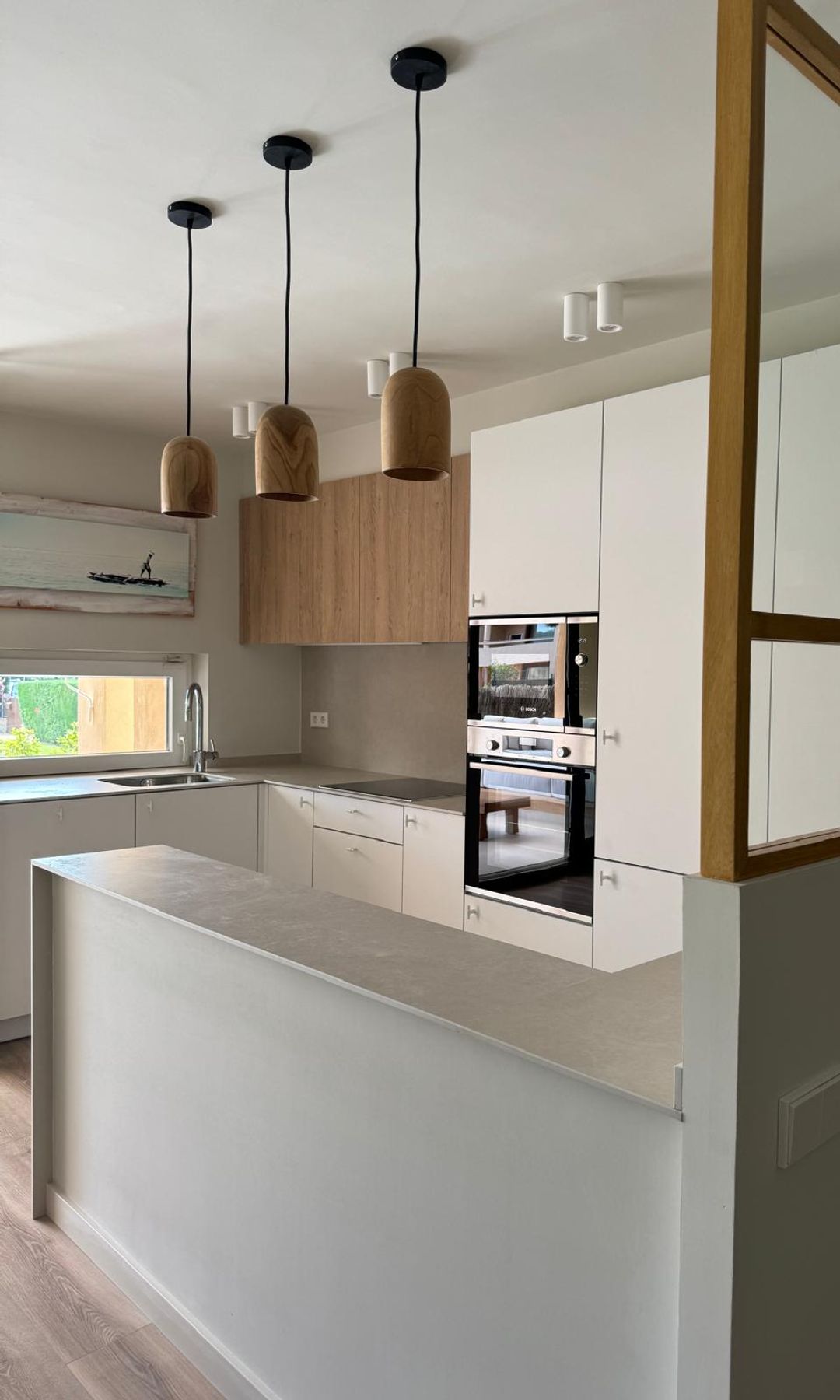 Cocina blanca, abierta con tabique de cristal con cuarterones de madera, focos empotrados y lámparas de techo