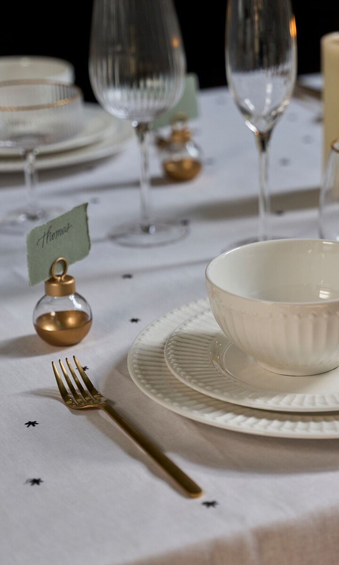 Mesa de Navidad con mantel blanco con estrellas, vajilla blanca, cristalería con el filo dorado, bola de Navidad como marcasitios