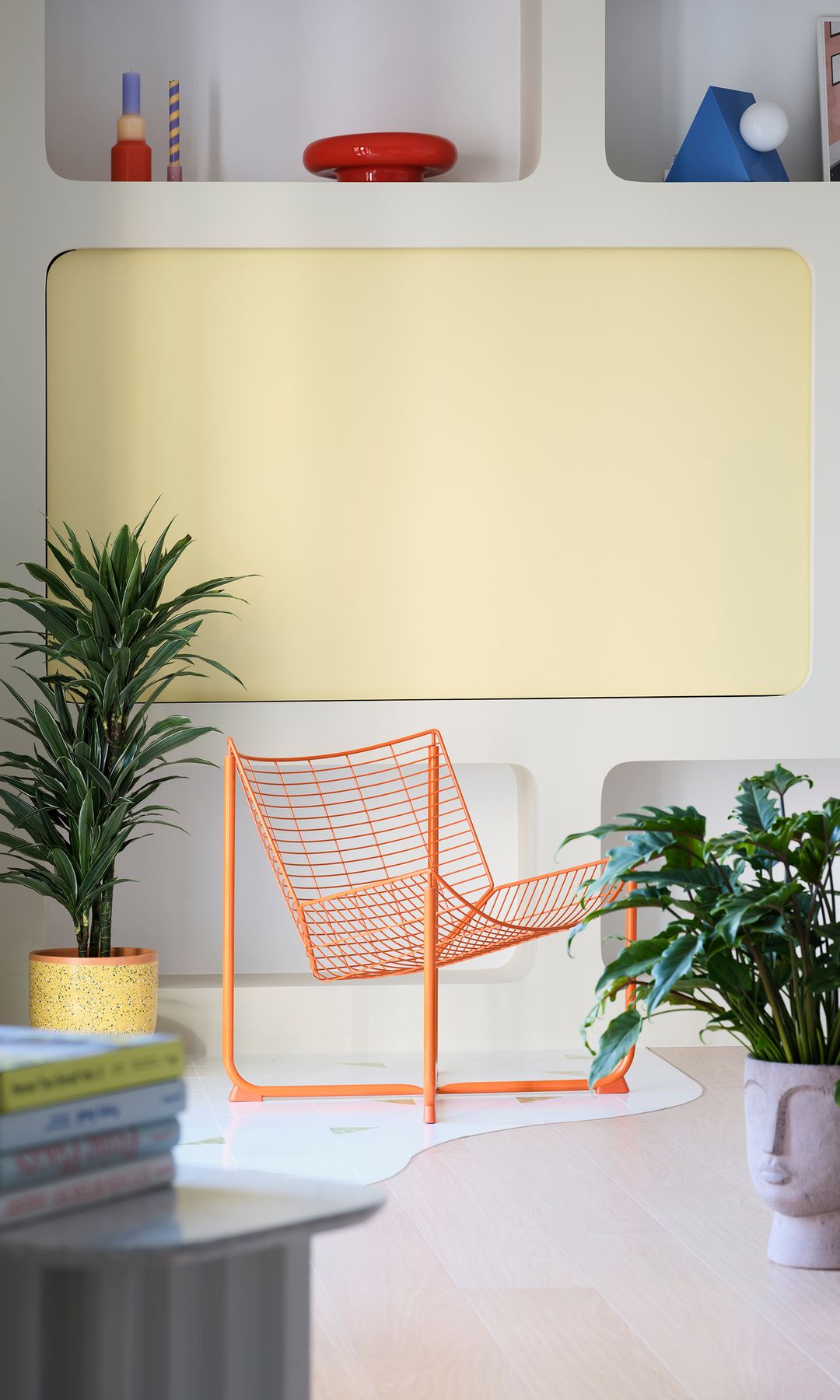 Salón con silla metálica de color naranja, macetas con plantas, estantería con nichos, televisión oculta tras un panel amarillo, adornos 